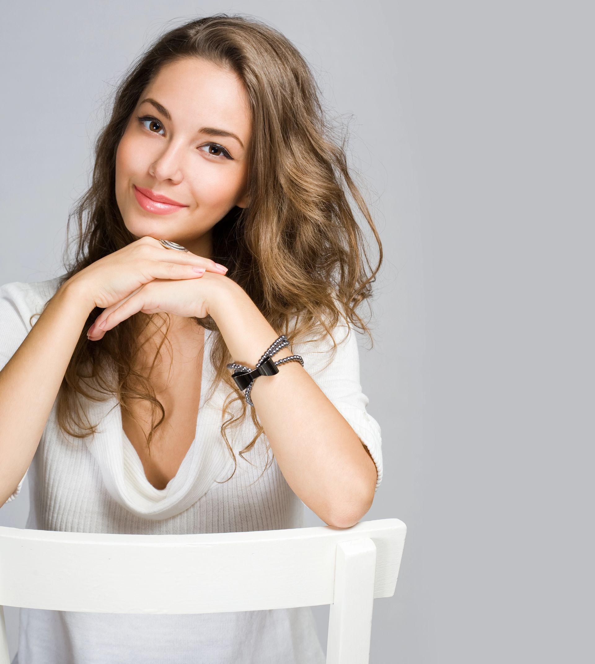 A woman is sitting in a white chair with her hands on her chin.