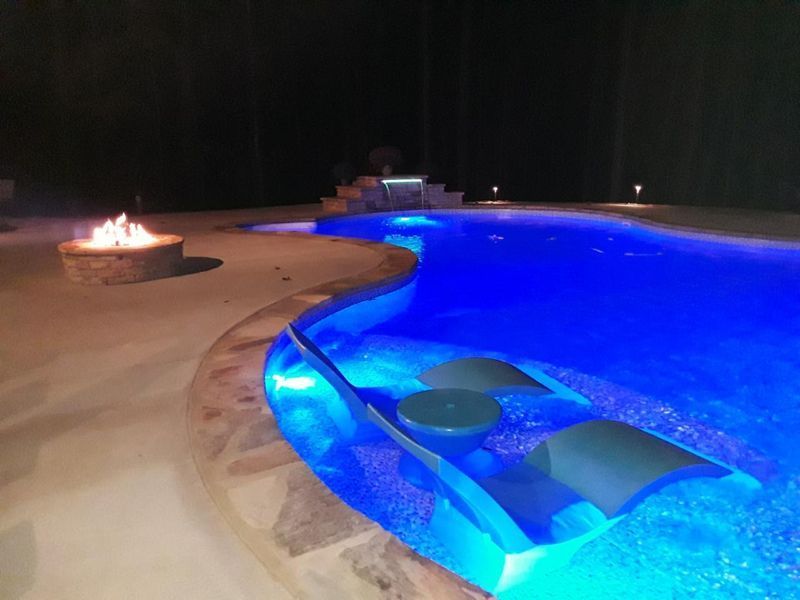 A large swimming pool is lit up at night with a fire pit in the background.