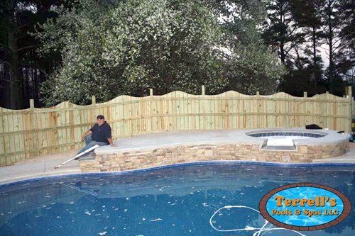 A man is sitting on the edge of a swimming pool next to a wooden fence.
