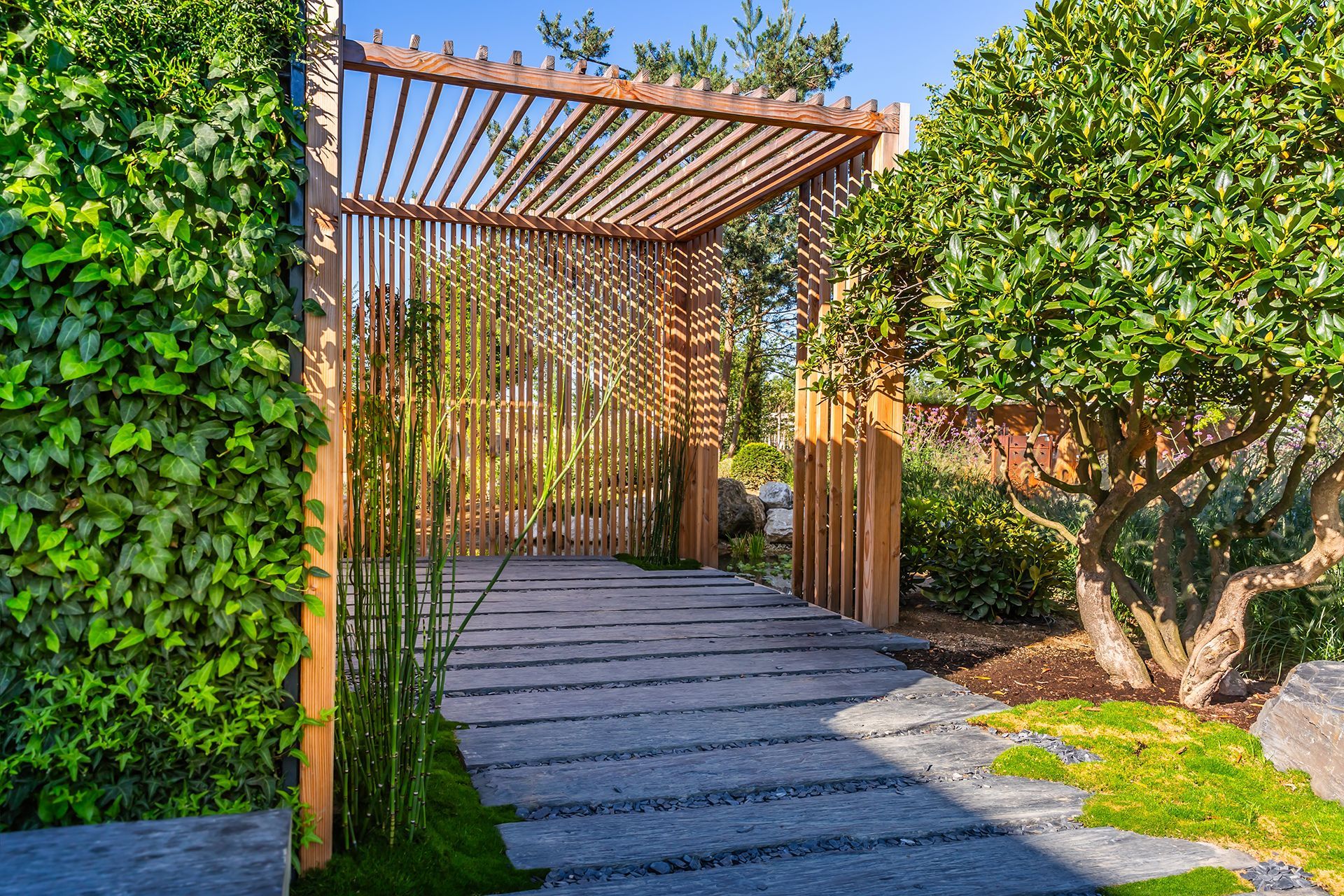 A wooden pergola over a stone walkway in a garden surrounded by trees.