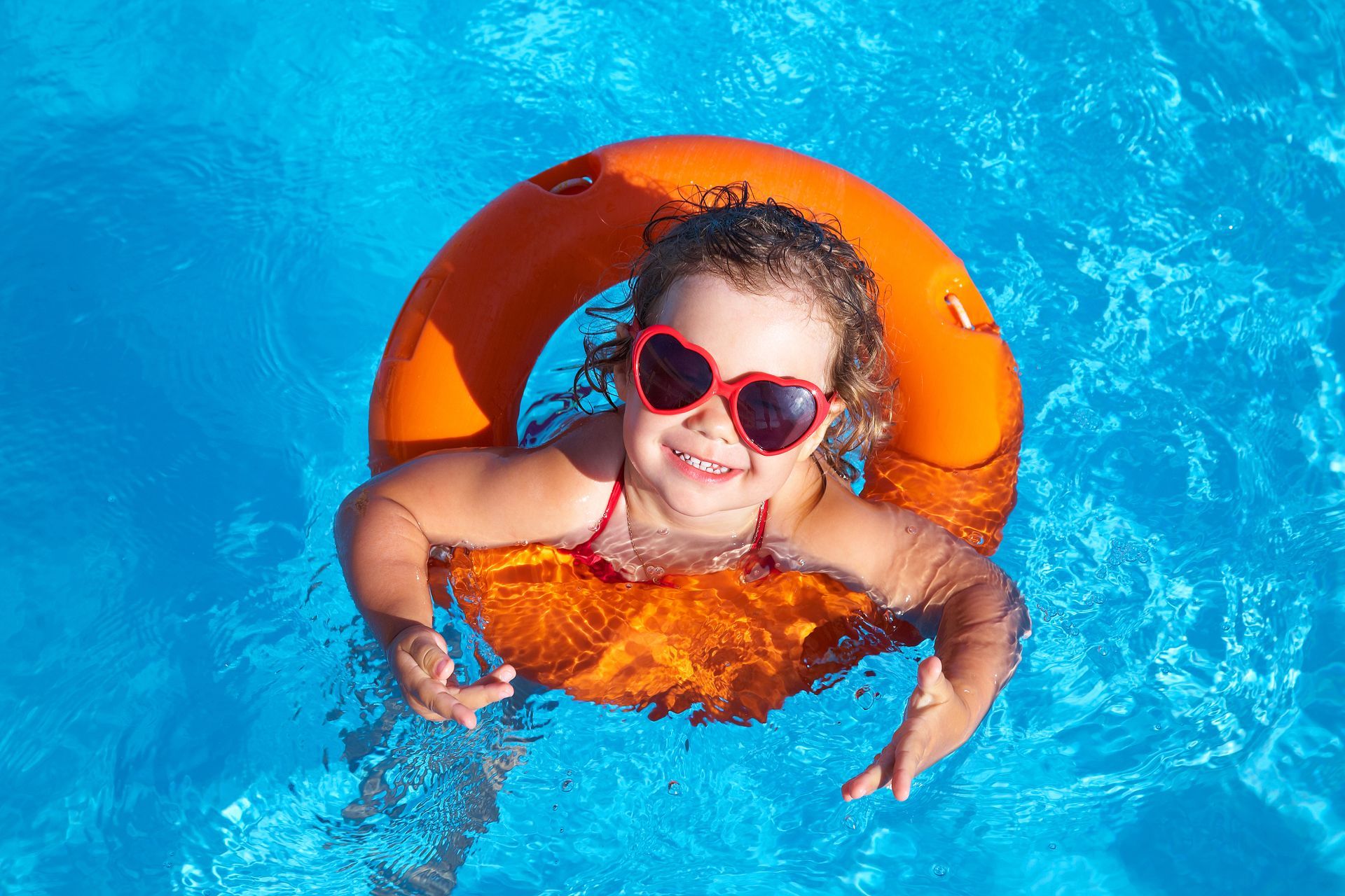 A little girl wearing sunglasses and an orange life preserver is swimming in a pool.