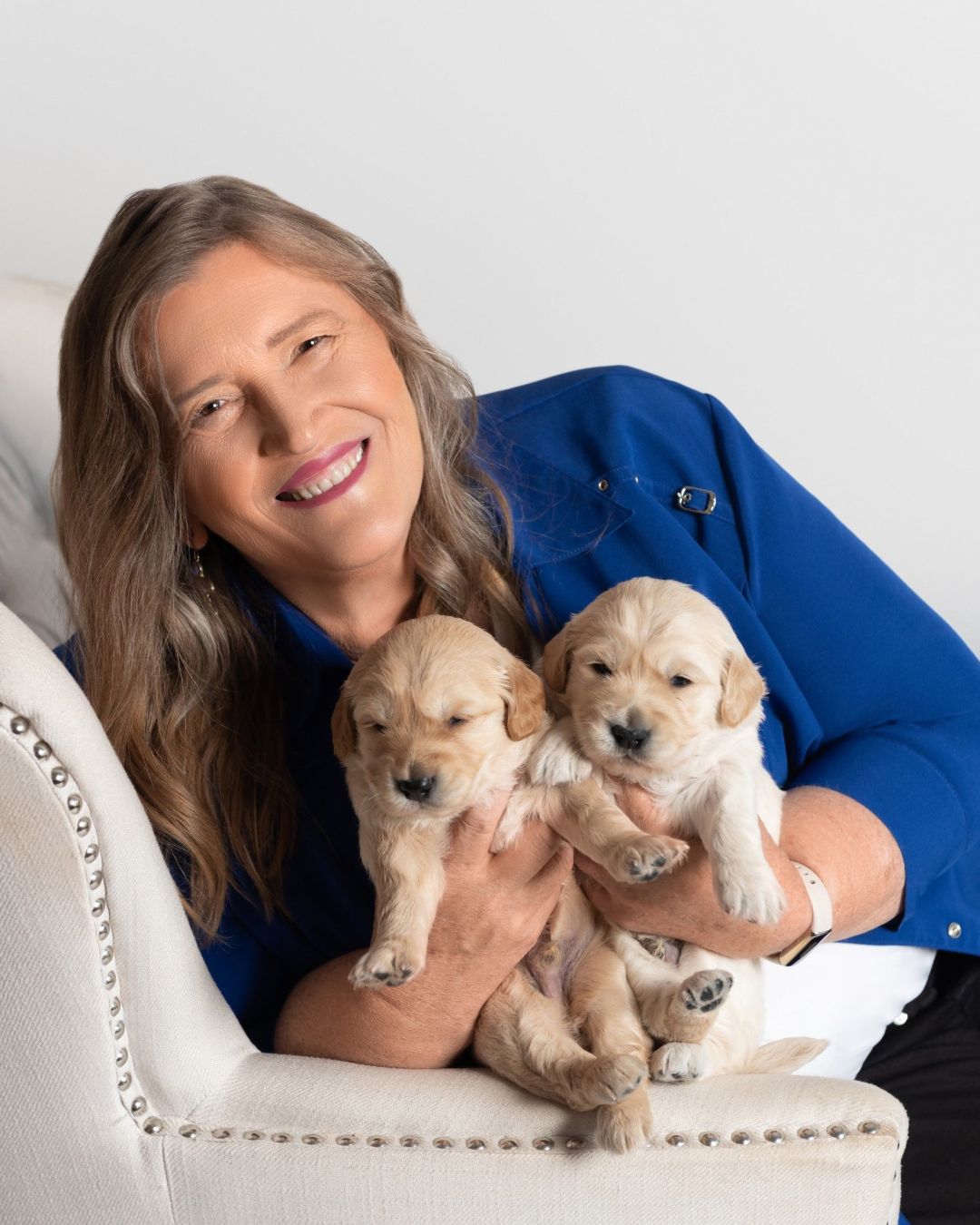 Kathy Burgess wearing a blue shirt is sitting on a light-colored chair, smiling while holding two Mini Golden Retriever puppies in her arms.