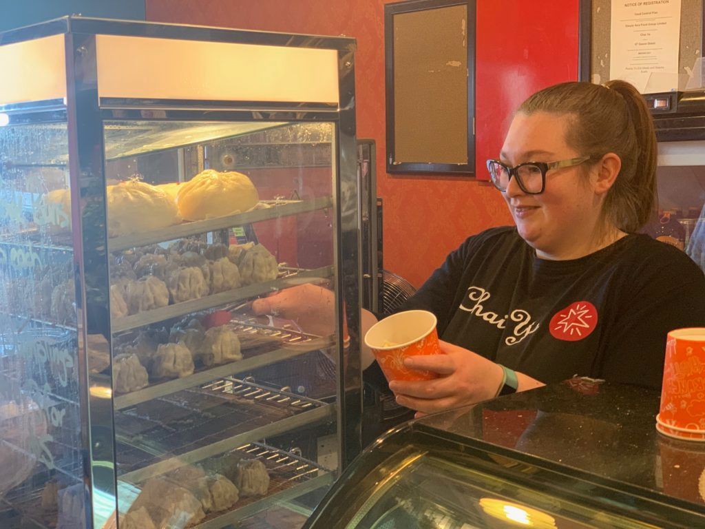 A woman is standing behind a counter in a bakery holding a cup of coffee.