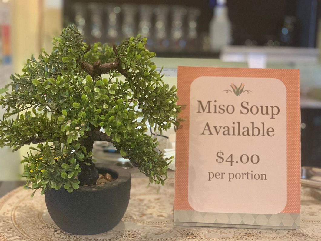 A small bonsai tree is sitting next to a sign that says miso soup available $ 4.00 per portion.