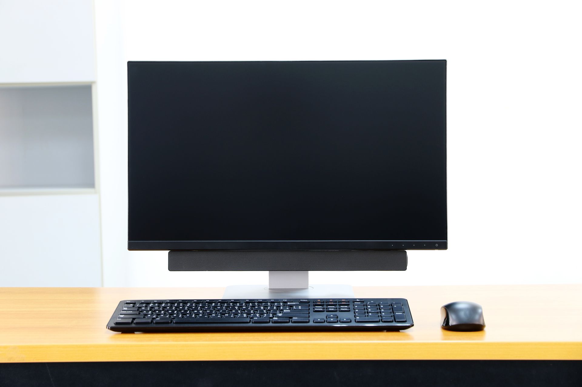 Computer setup on wooden desk: black monitor, keyboard, mouse. White wall in background.