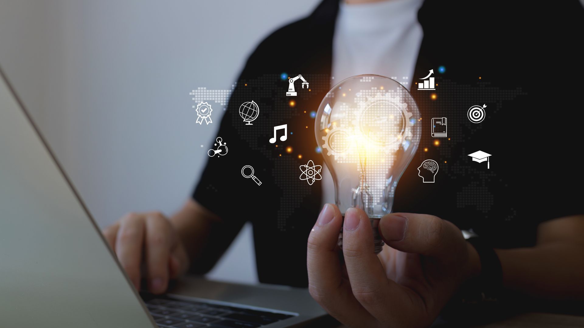 Person holding a lit lightbulb, with icons floating around it, while using a laptop.