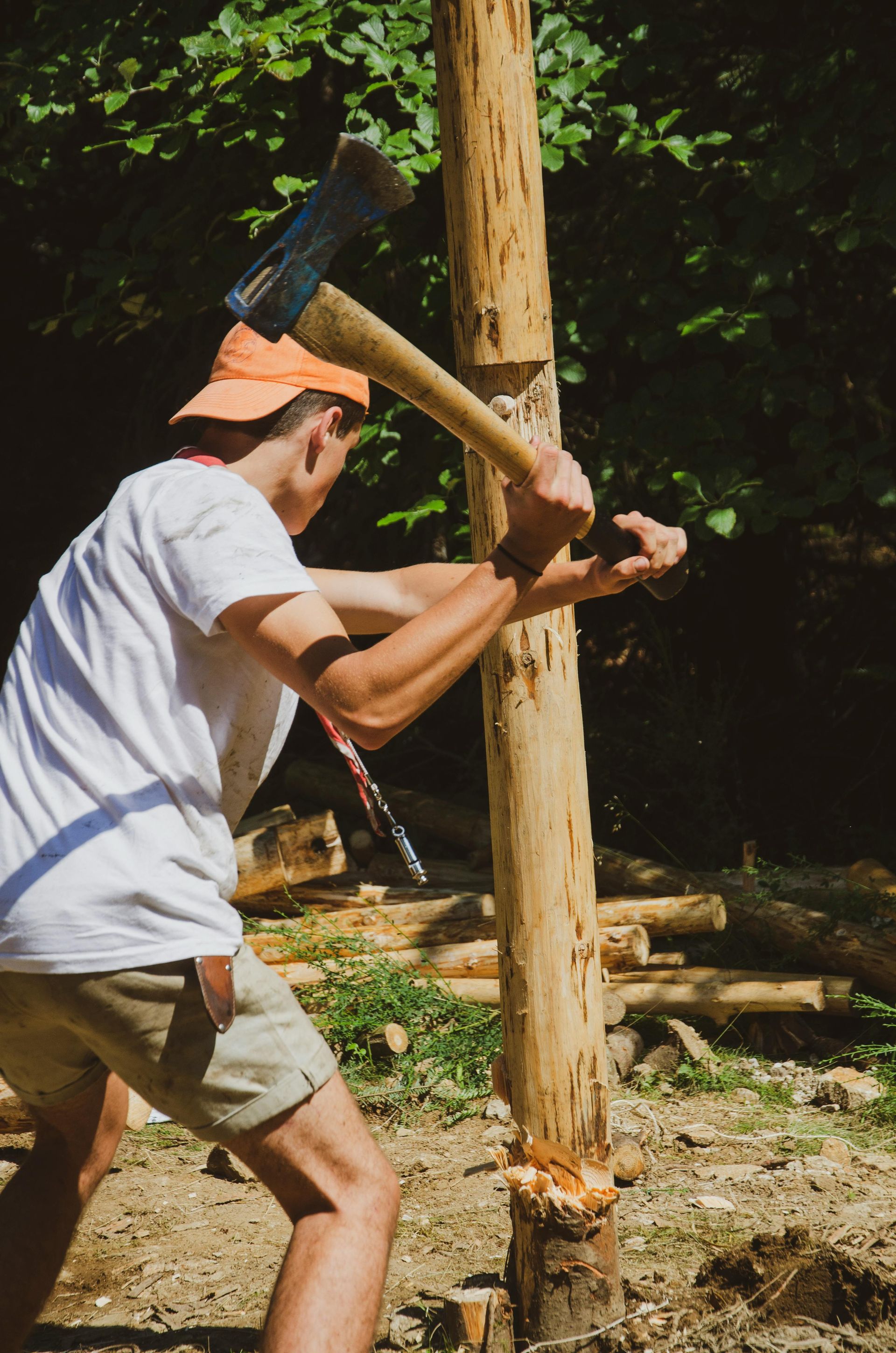Person chopping wood with an ax. Wearing a white shirt, orange hat, and khaki shorts, outside.