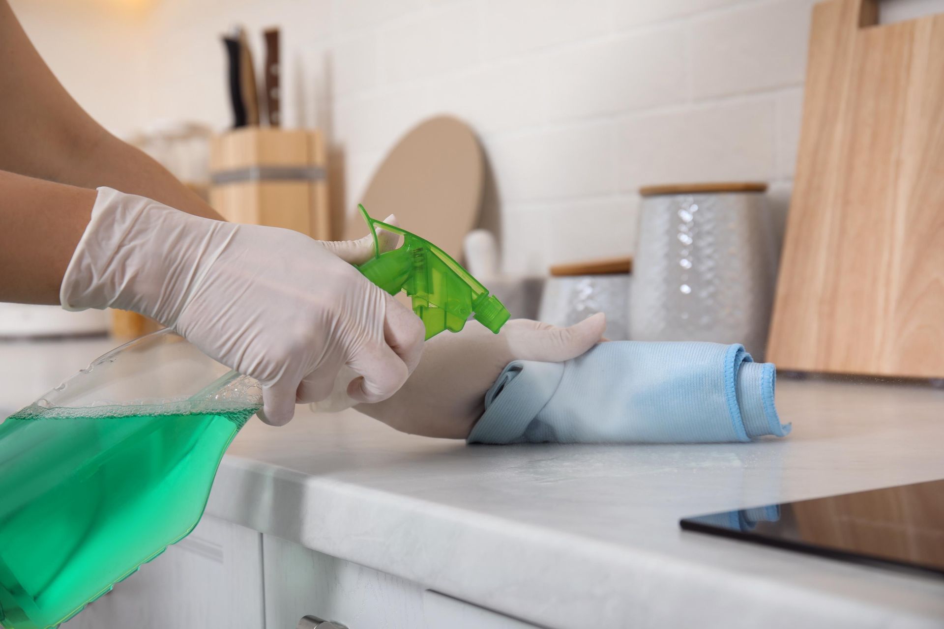 Hands in white gloves spraying green cleaning solution onto a light blue cloth on a kitchen counter.
