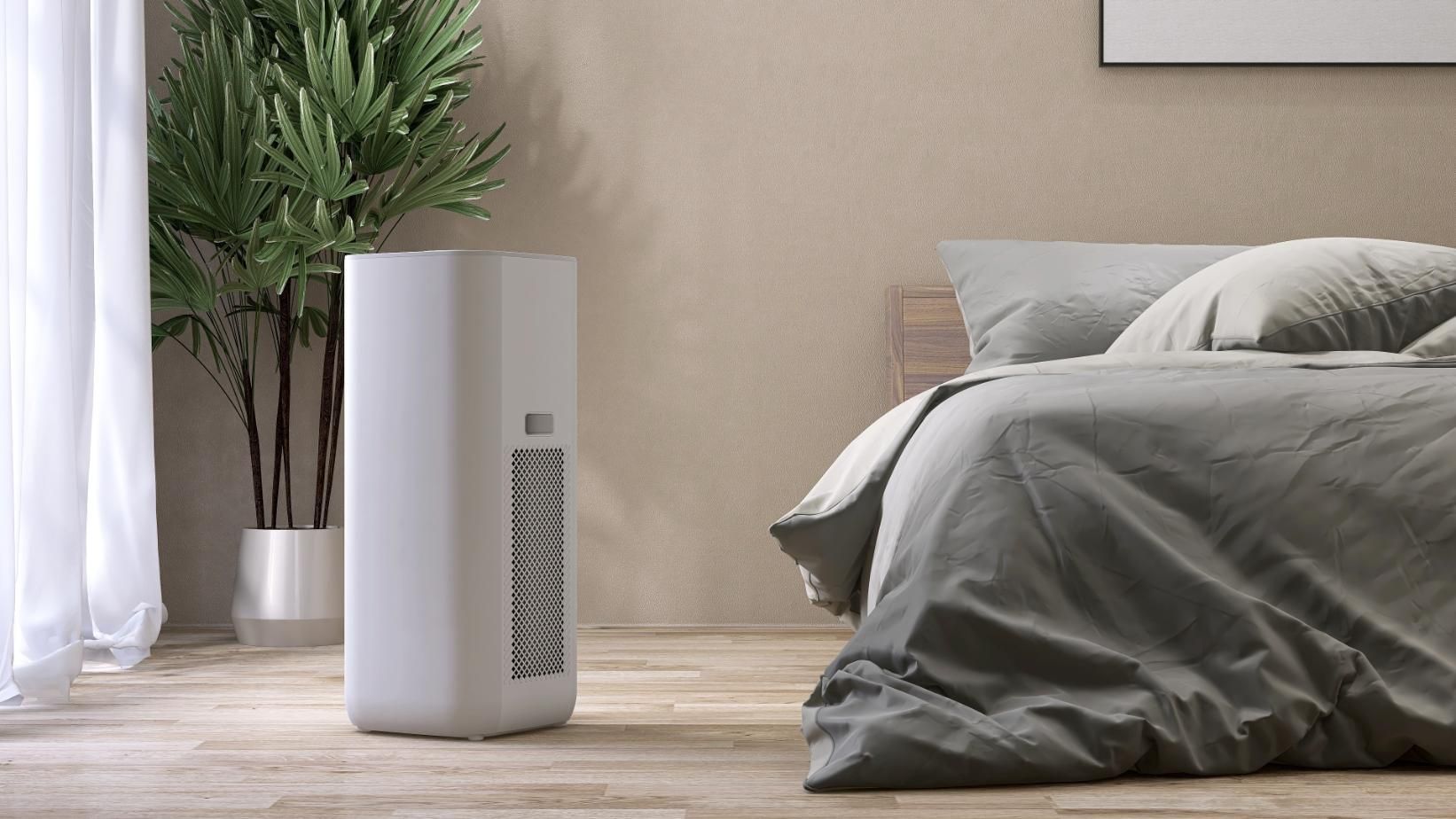 A white air purifier stands on a light wood floor in a bedroom.