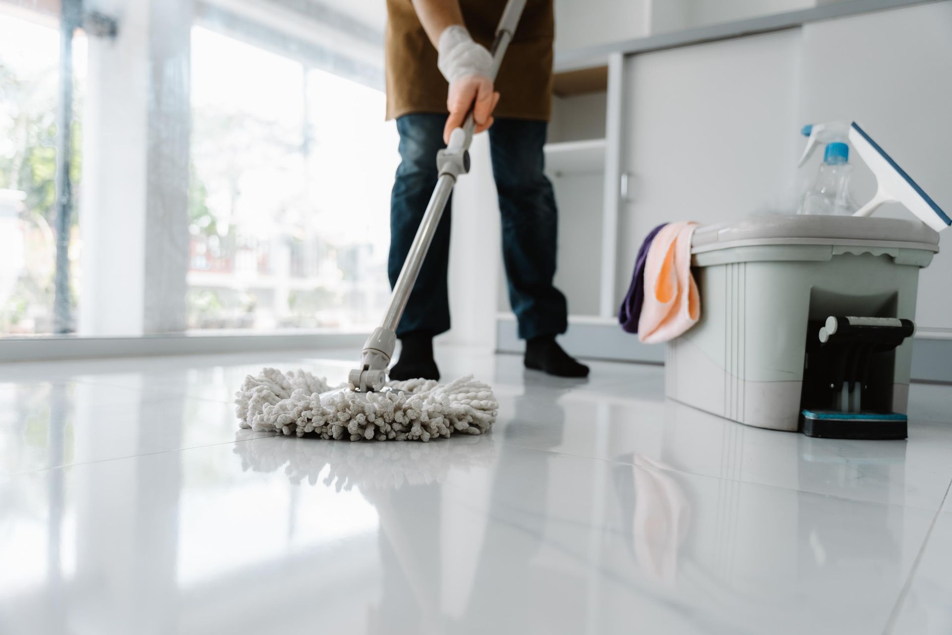 A person mops a white tiled floor in a brightly lit room next to a bucket with cleaning supplies.