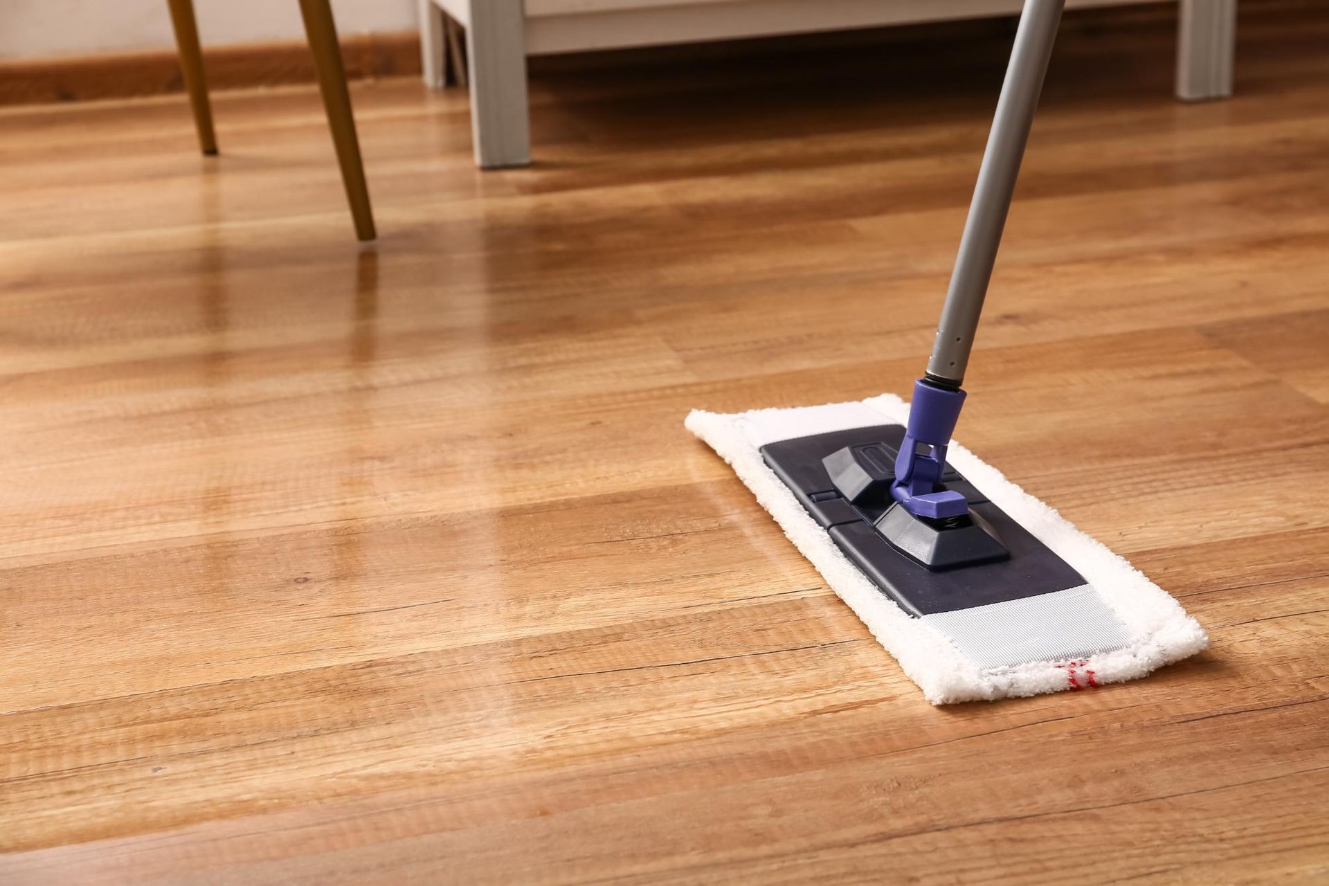 A flat mop with a white microfiber pad cleans a wooden floor in a room.