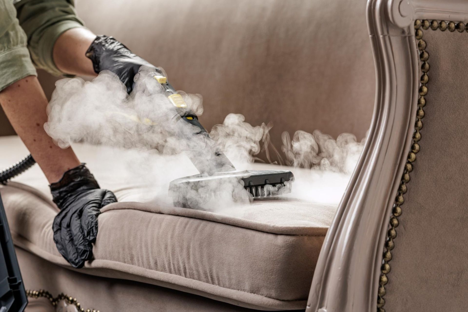 A person wearing black gloves uses a steam cleaner to sanitize a light-colored sofa.