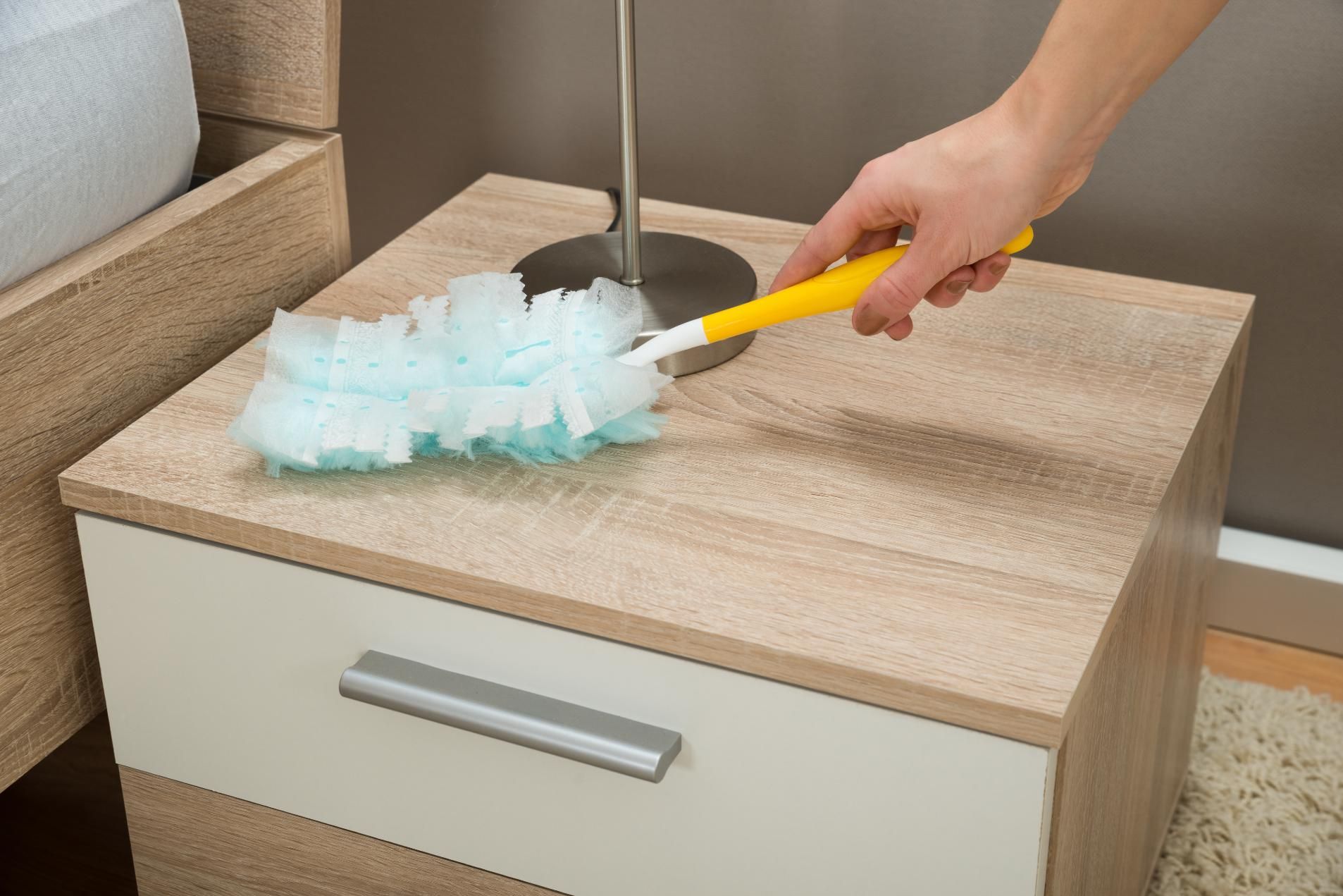 A hand uses a yellow-handled blue duster to clean the top of a wooden bedside table next to a lamp.