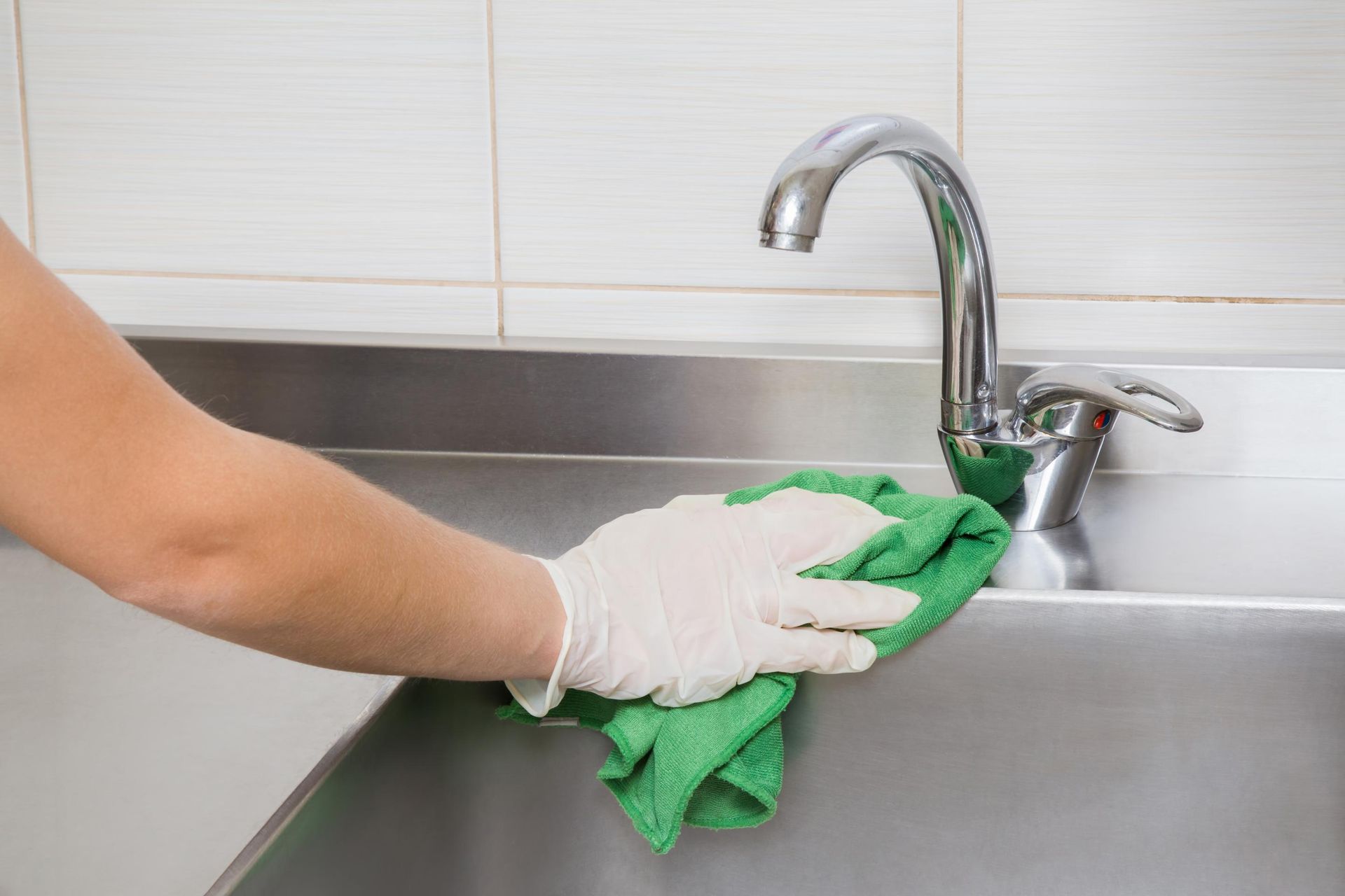 A person wearing a white glove wipes a stainless steel kitchen sink faucet with a green cloth.