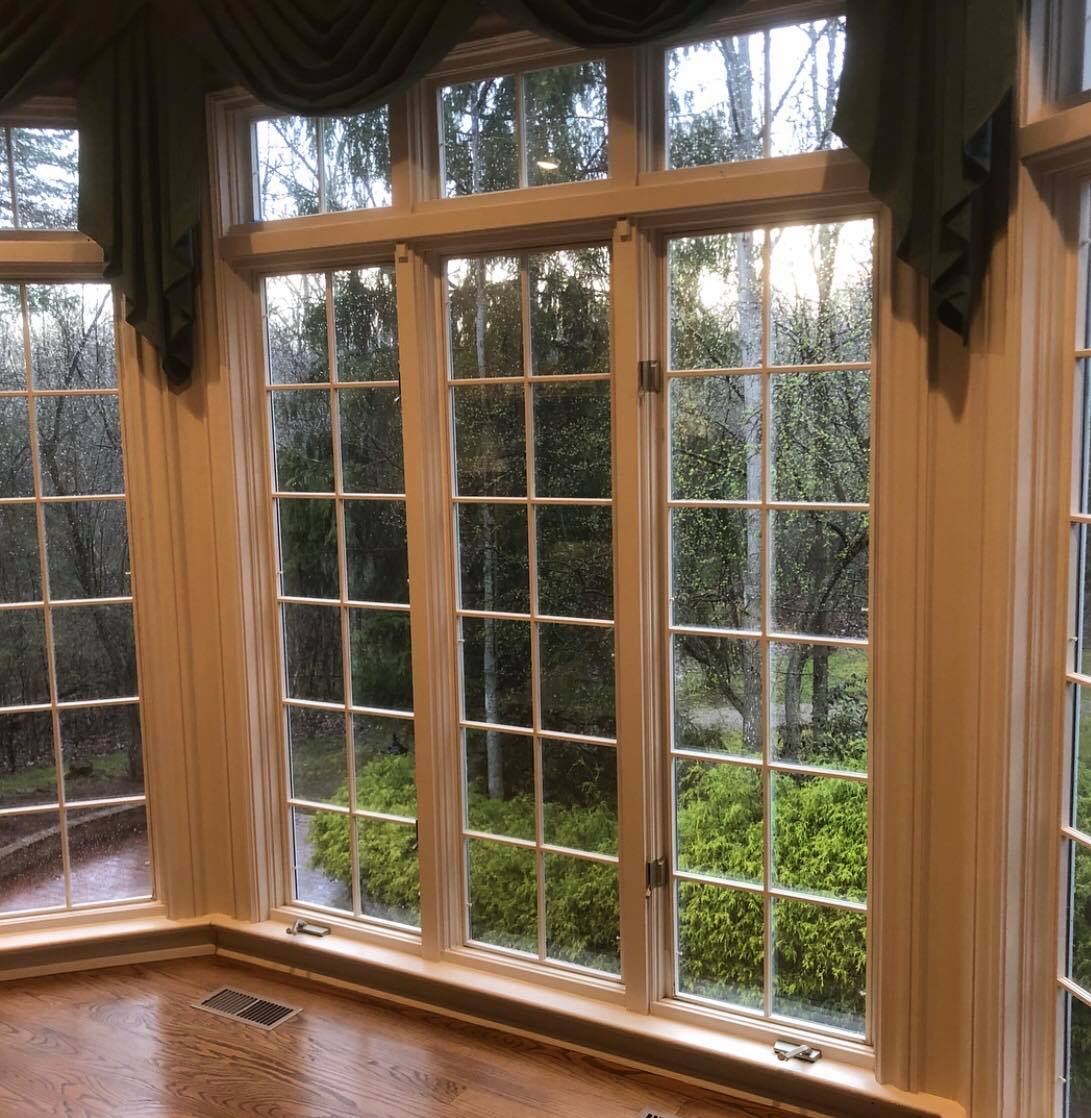 White-framed window corner with multiple panes overlooking a blurry, green, outdoor scene. Green valance. Wooden floor.
