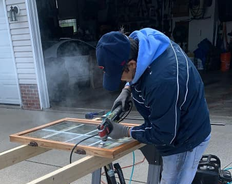 Person using a power tool to cut a window pane outdoors. Gray gloves, blue jacket, cap, and a wooden frame visible.