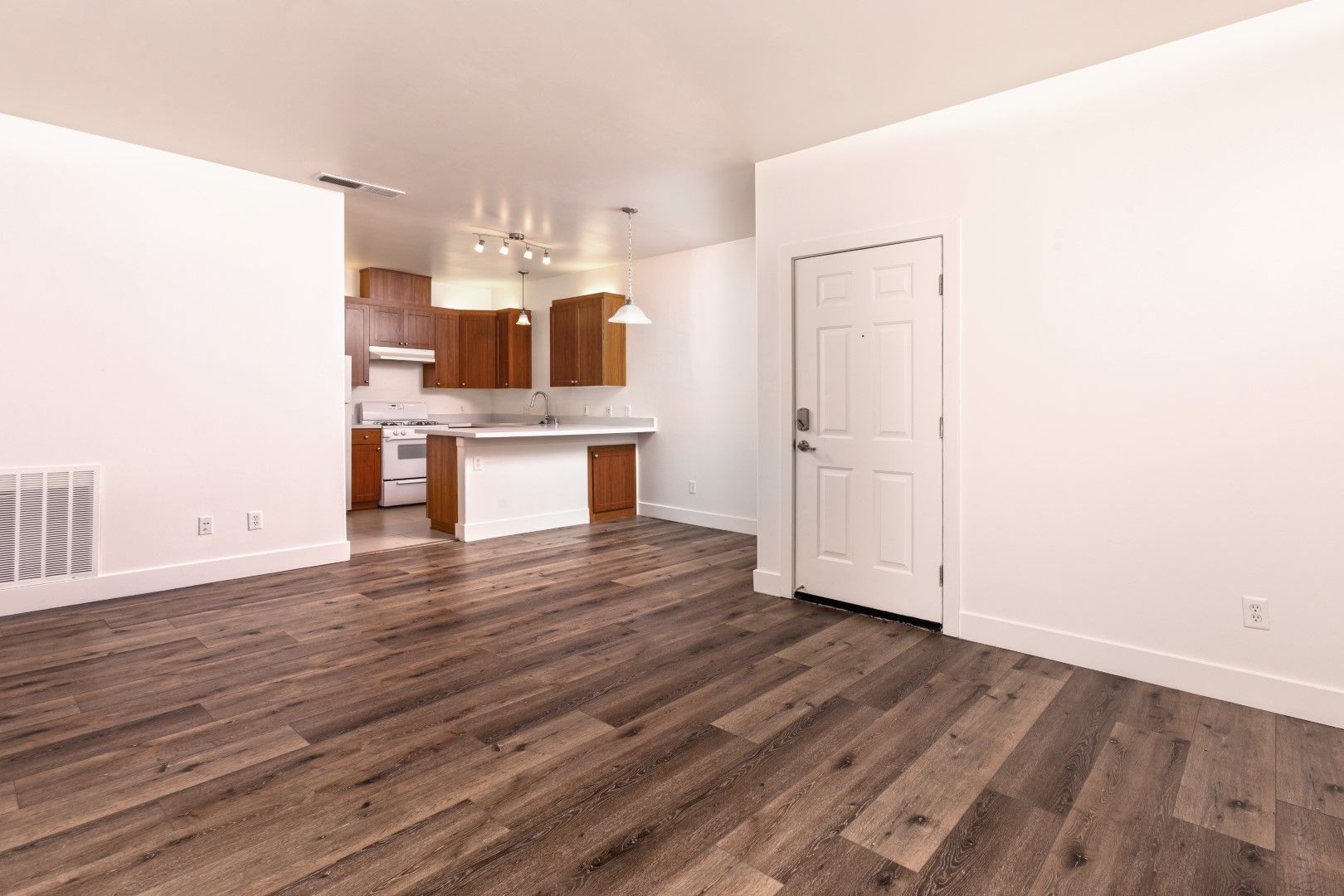 An empty living room with hardwood floors and a kitchen in the background.