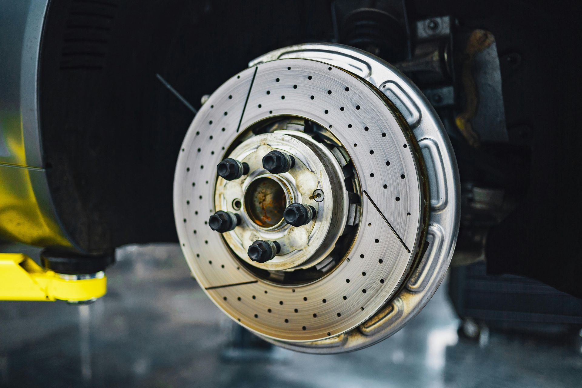 Car brake rotor, silver with drilled holes and slotted grooves, mounted on vehicle.