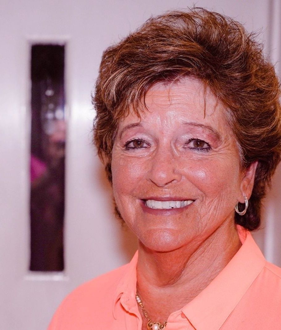 Smiling older woman with short brown hair in a peach shirt, standing indoors.