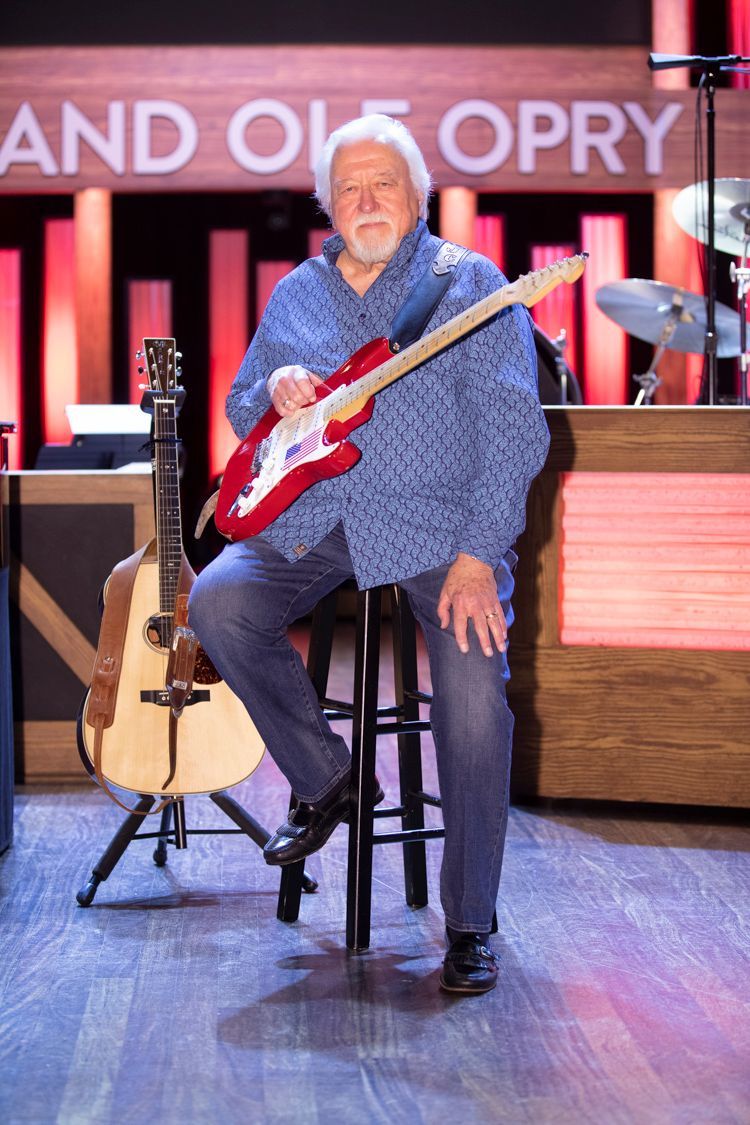 Man seated, holding a red electric guitar at the Grand Ole Opry stage; acoustic guitar nearby.