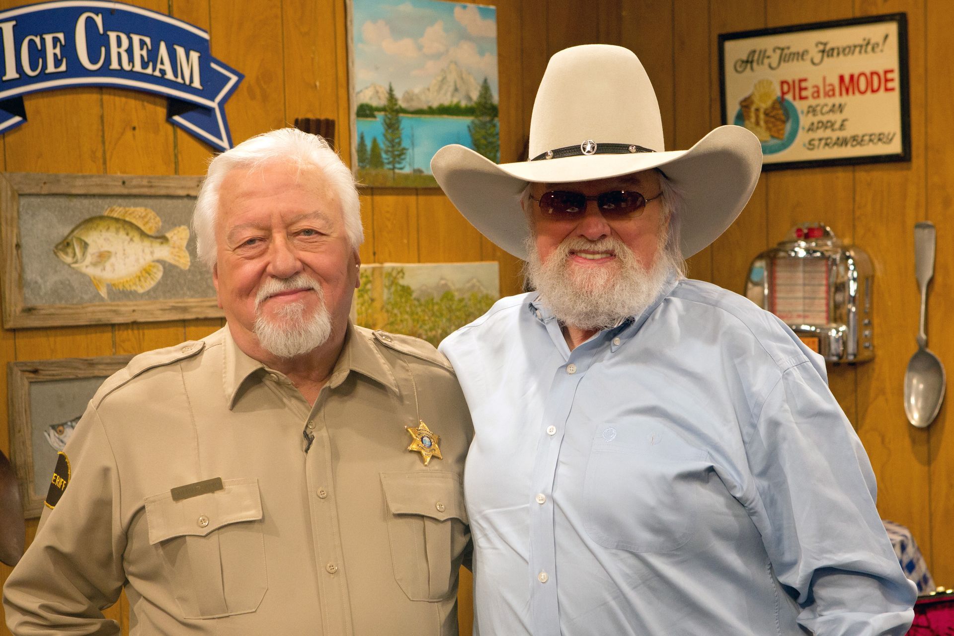 Two older men posing together indoors; one in a sheriff's uniform, the other in a cowboy hat and light blue shirt.