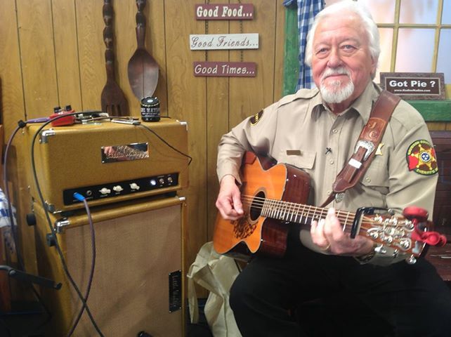 Man in uniform plays acoustic guitar next to a guitar amp in a rustic setting.