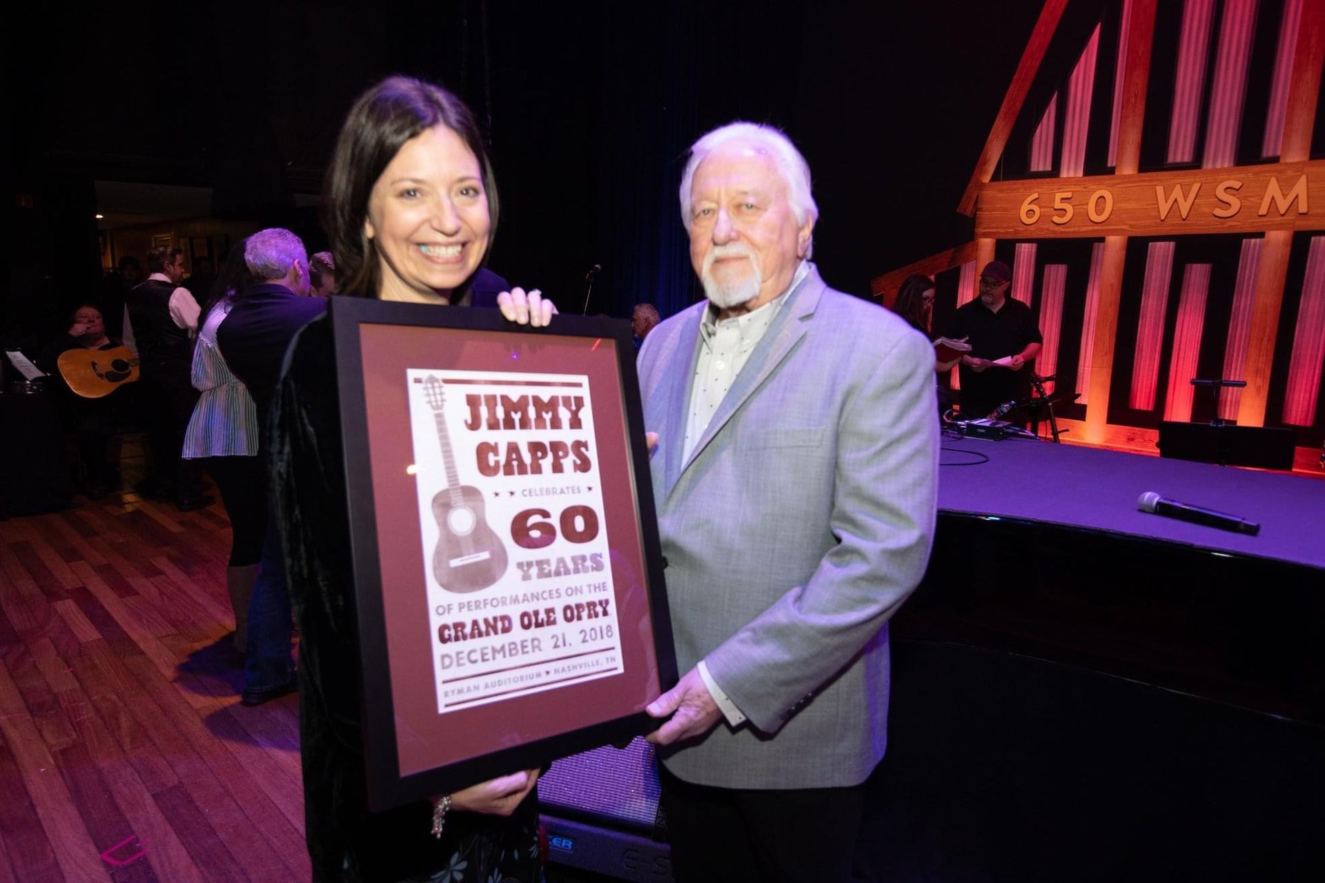 Woman and man hold a framed award onstage. The award is for Jimmy Capps' 60 years on the Grand Ole Opry.