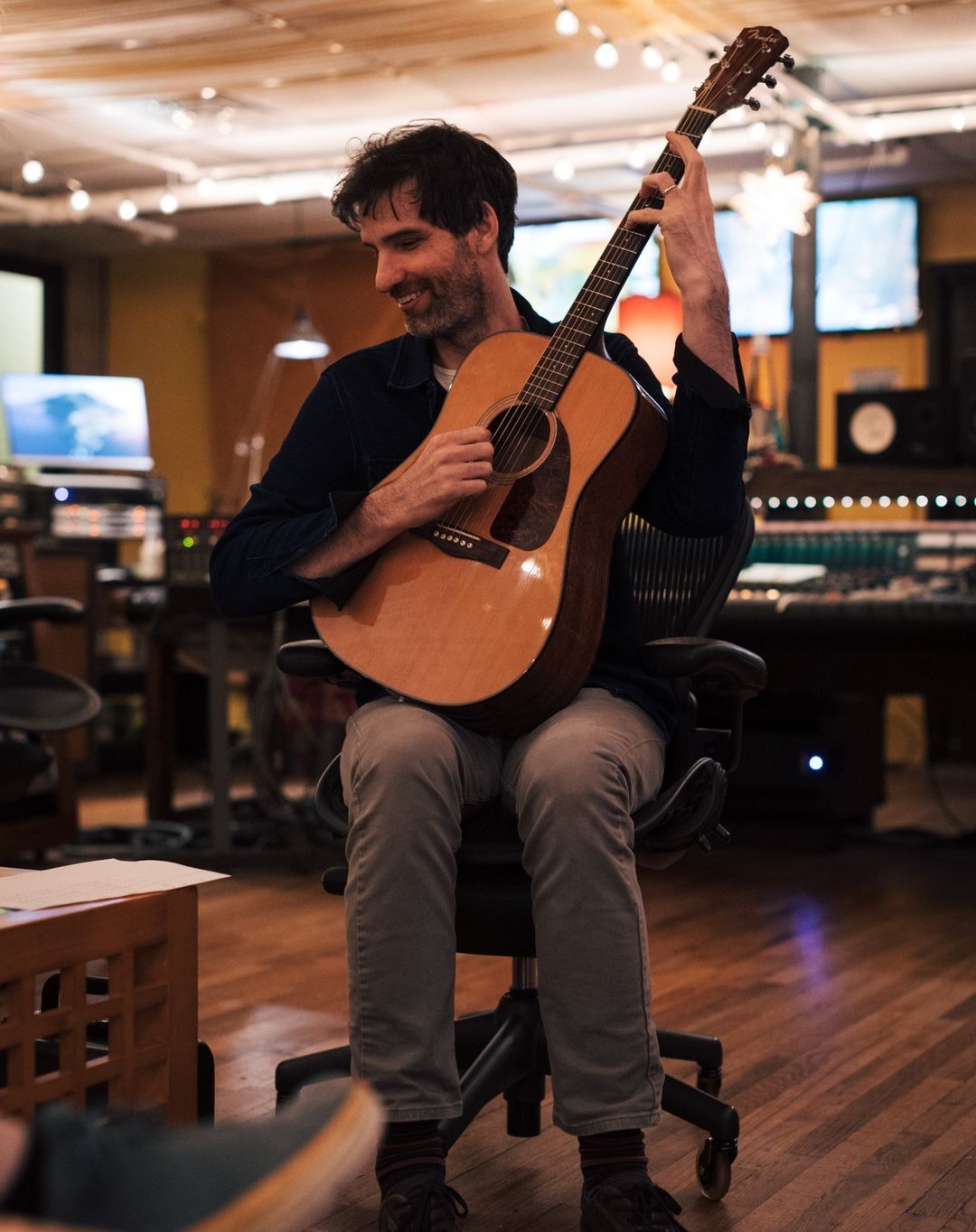 Man seated, playing acoustic guitar; in recording studio. He's smiling, wearing casual clothes.