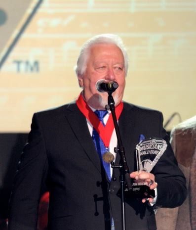 Man in a suit holding an award, speaking into a microphone. He wears a medal, stands in front of a backdrop.