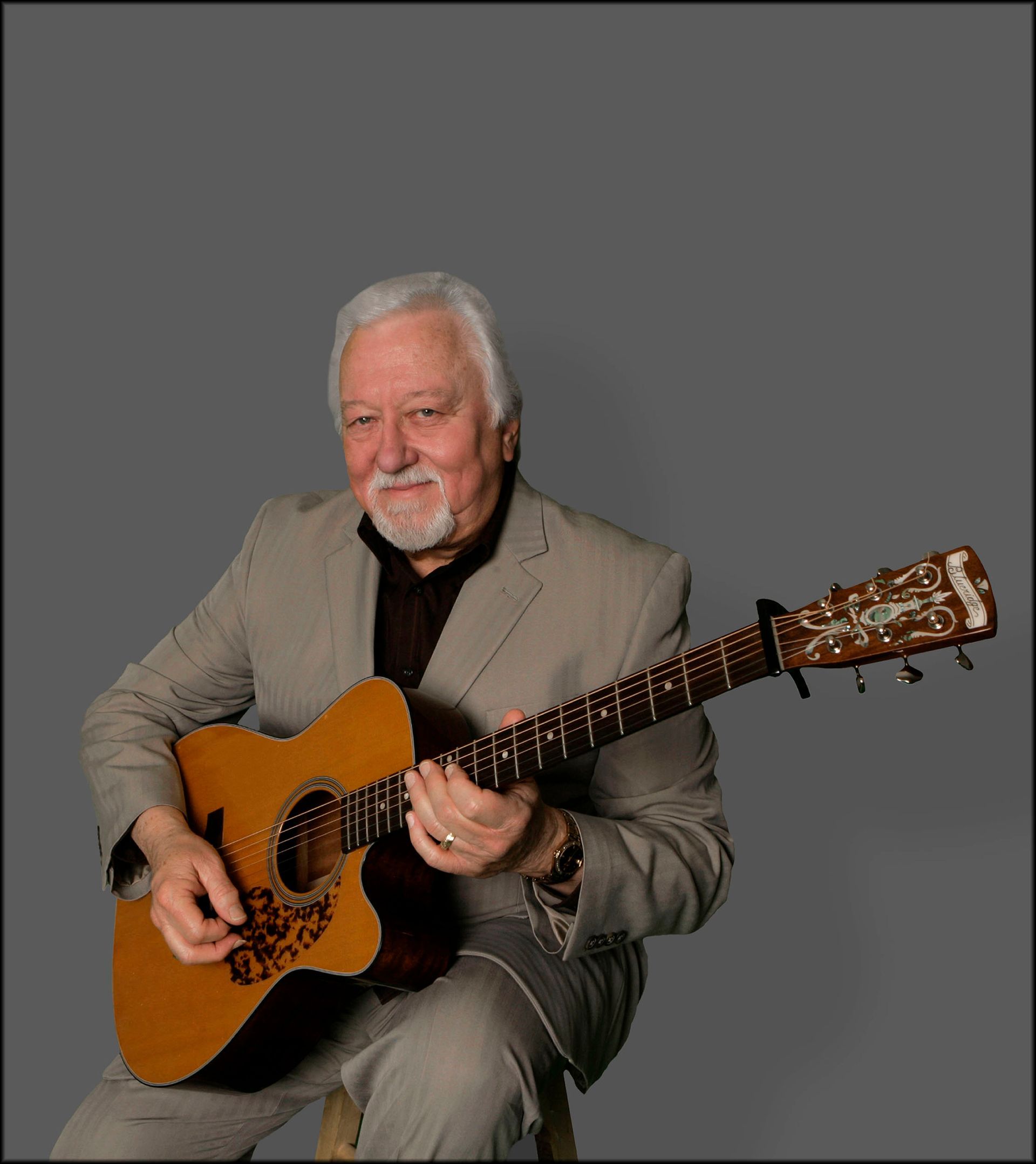 Man seated, playing acoustic guitar. He wears a suit. Dark background.