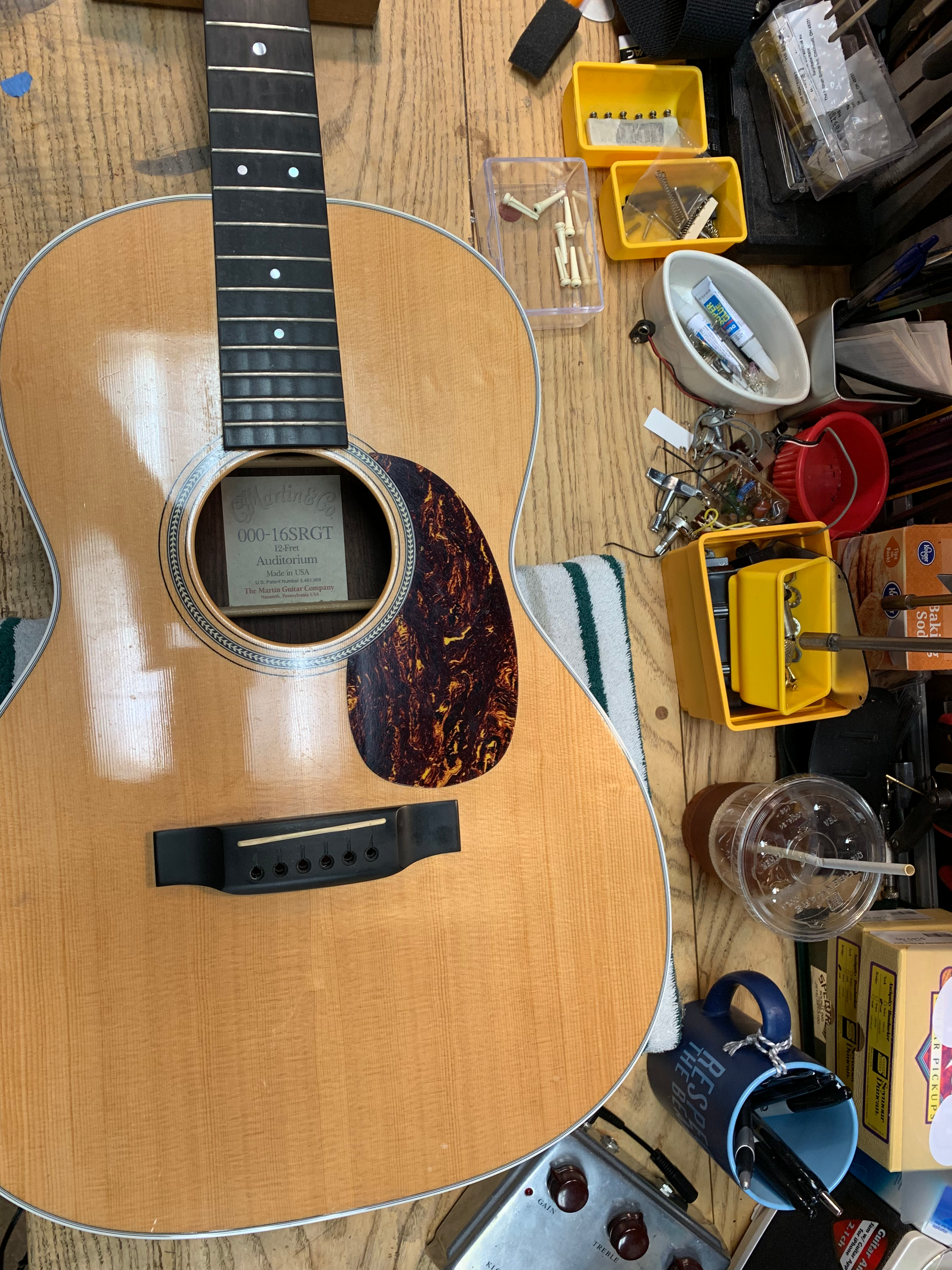 Acoustic guitar on a workbench. Natural wood finish, tortoiseshell pickguard, surrounded by tools and parts.