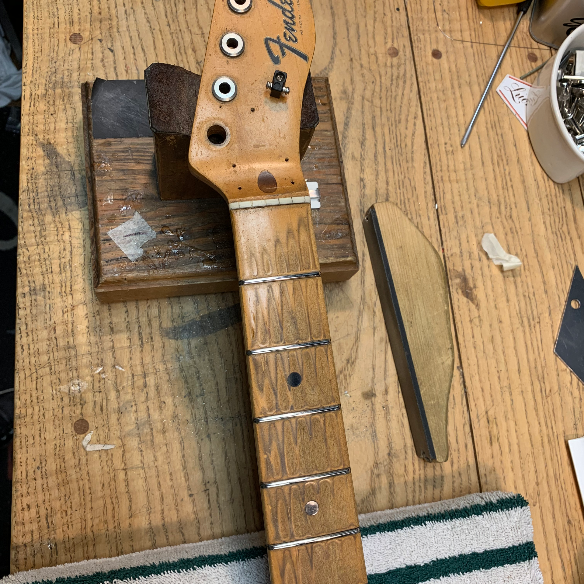 Guitar neck being repaired on a workbench. Shows frets, headstock, and wooden sanding tools.