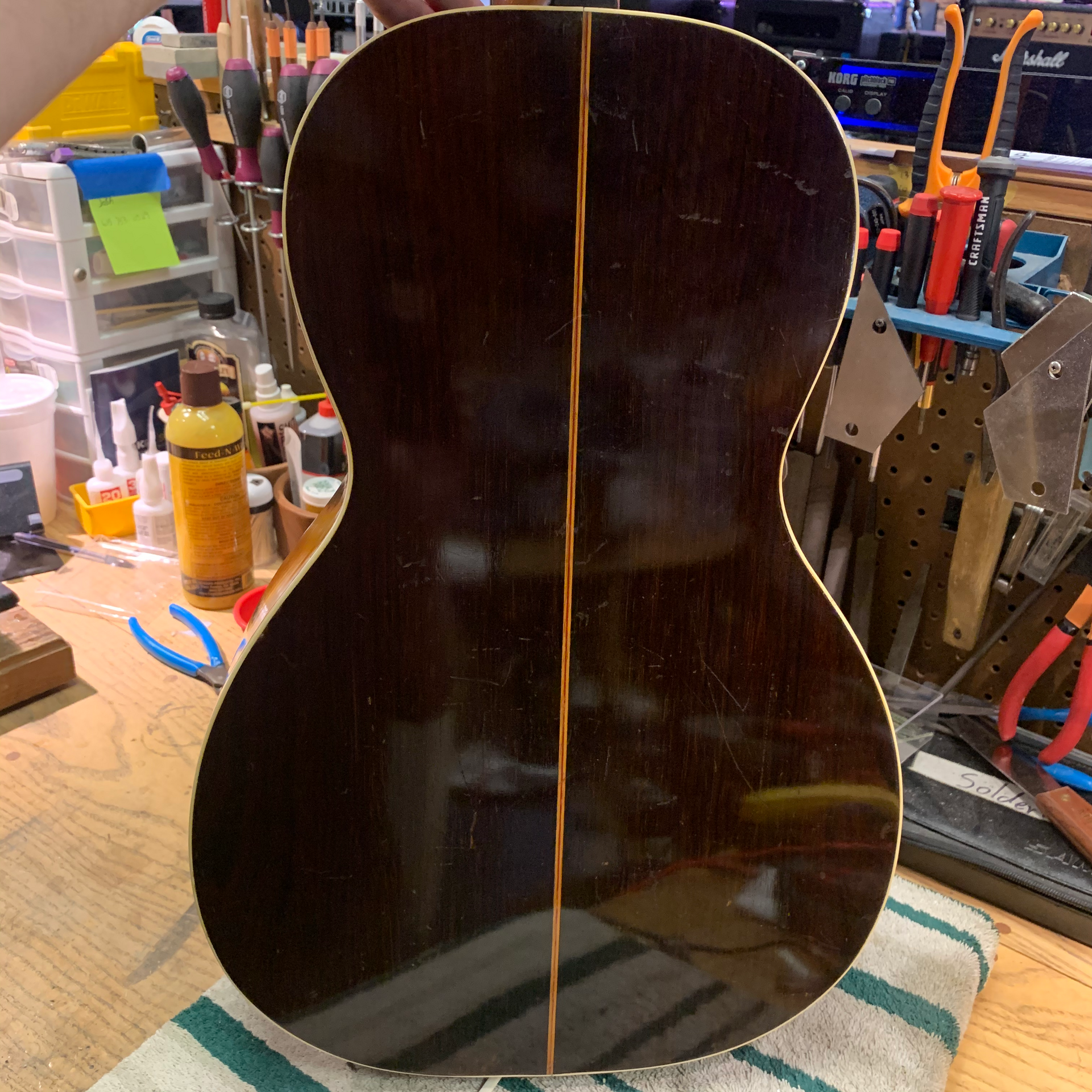Dark acoustic guitar back, with center strip. Surrounded by a repair shop setting with tools and equipment.