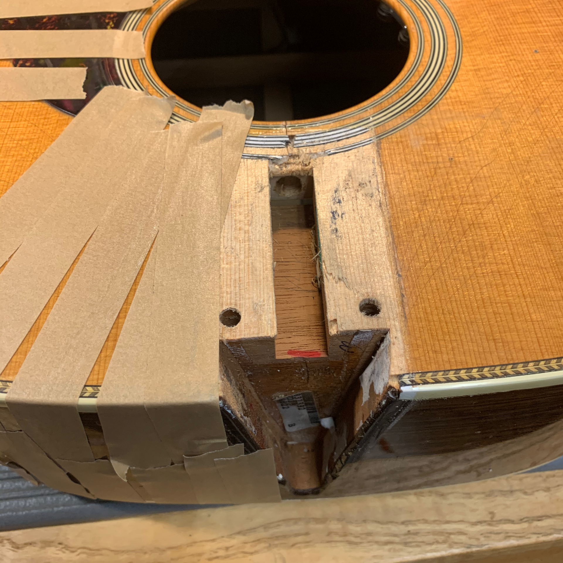 Guitar body, neck joint partially disassembled for repair. Tape protects the soundboard. Wood tones.