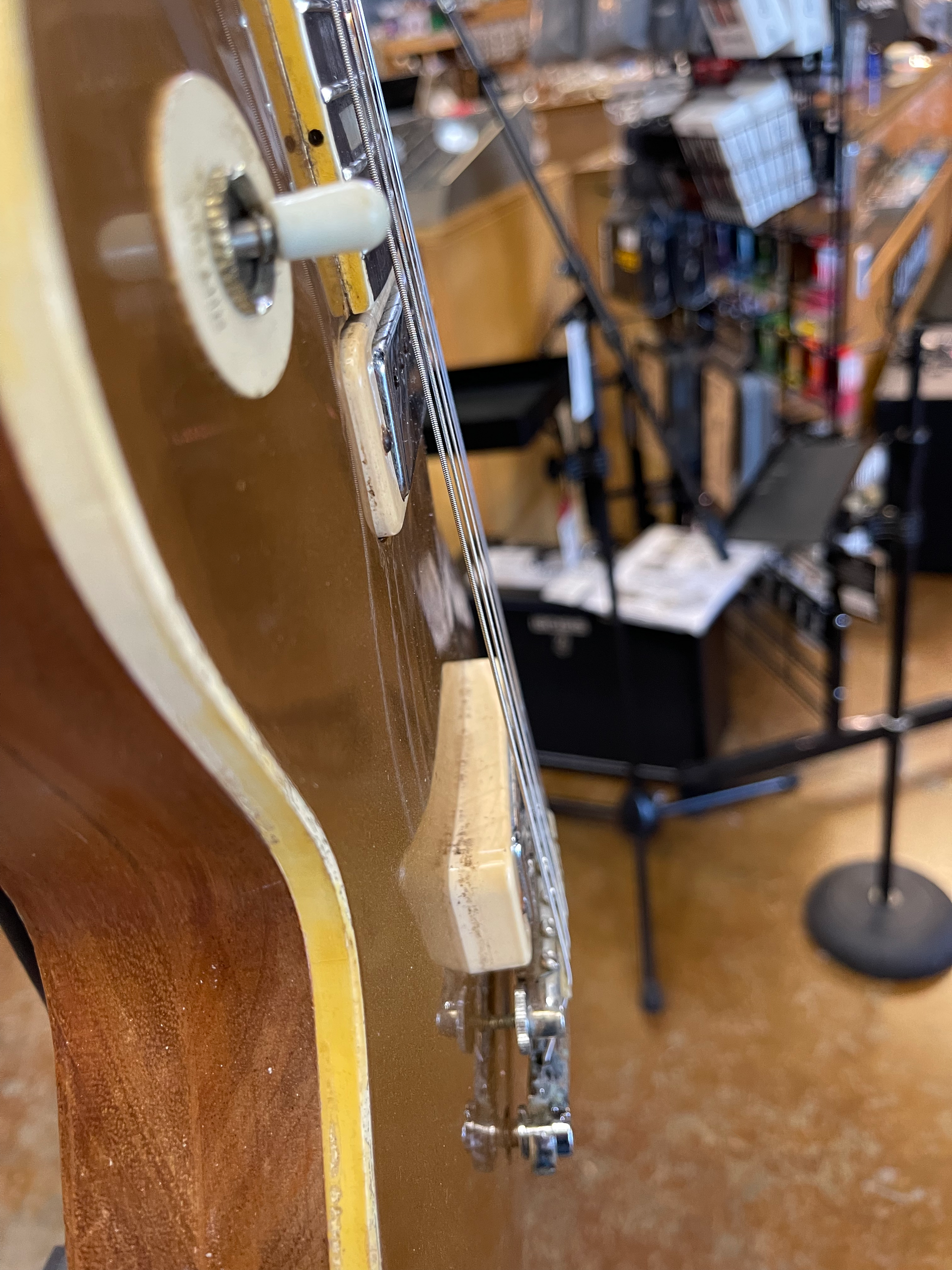 Close-up of a gold electric guitar. Side view showing fretboard, hardware, and cream binding. Blurred shop background.