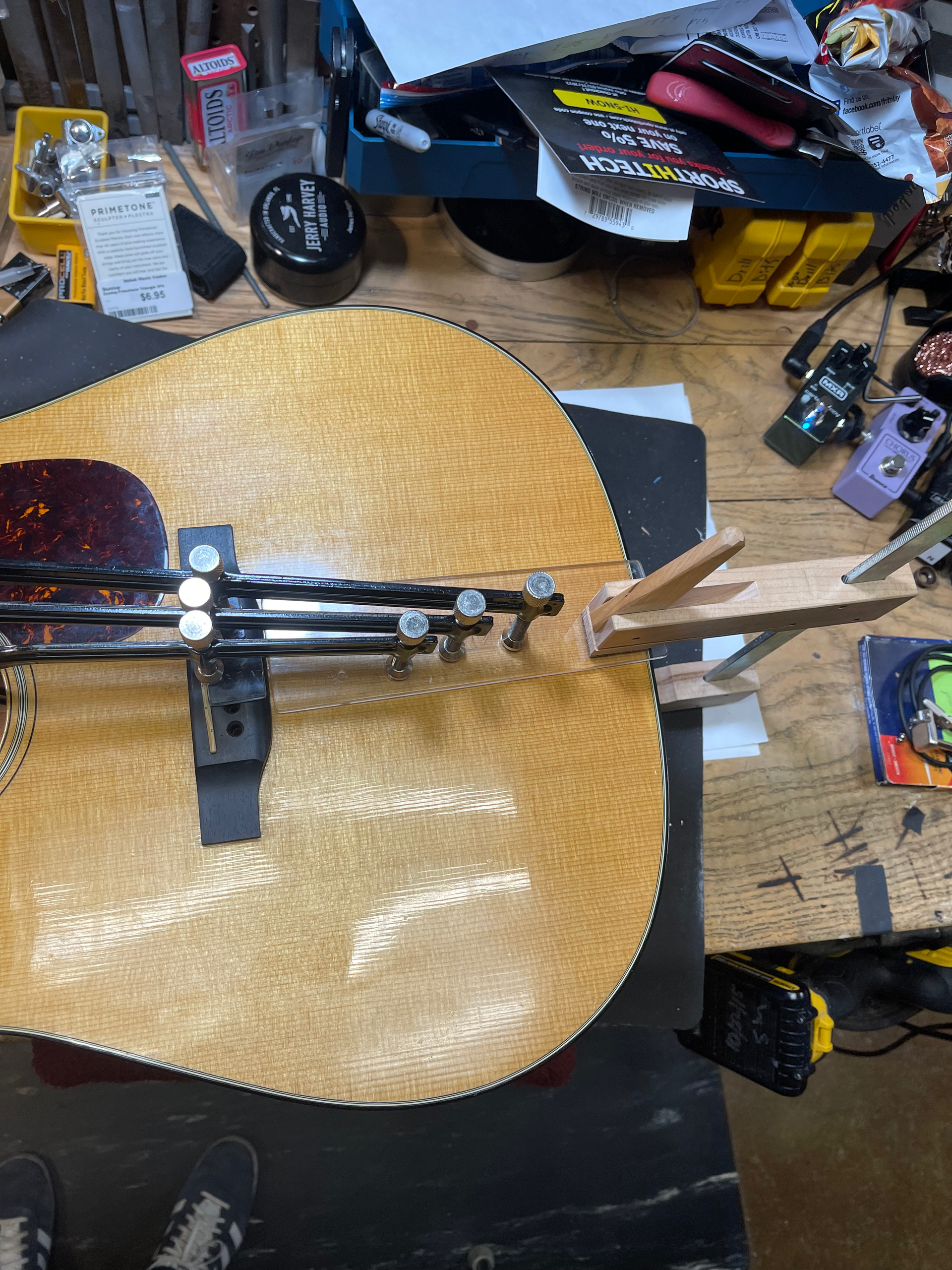 Acoustic guitar in a repair shop, clamped for neck work. Light wood grain, tools and parts visible.