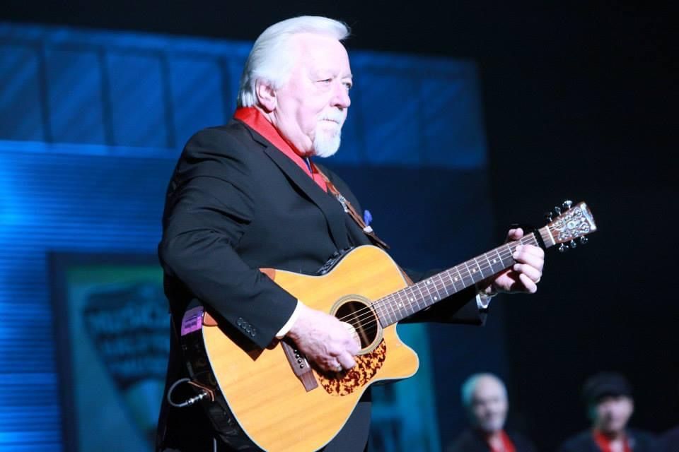 Man in black suit playing acoustic guitar on stage, holding a pick.