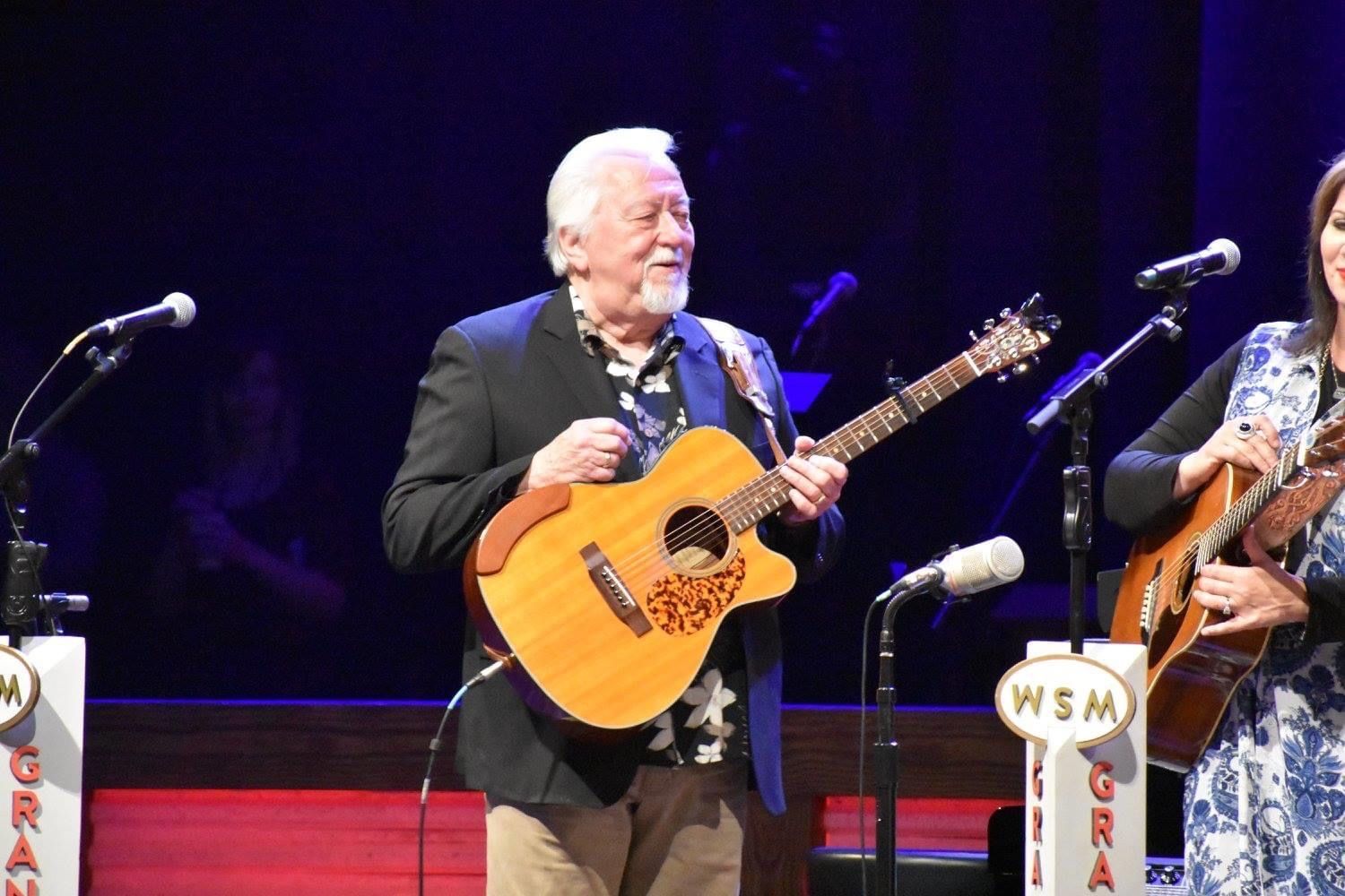 Man playing acoustic guitar on stage, smiles. Two others beside him with instruments, WSM logo visible.