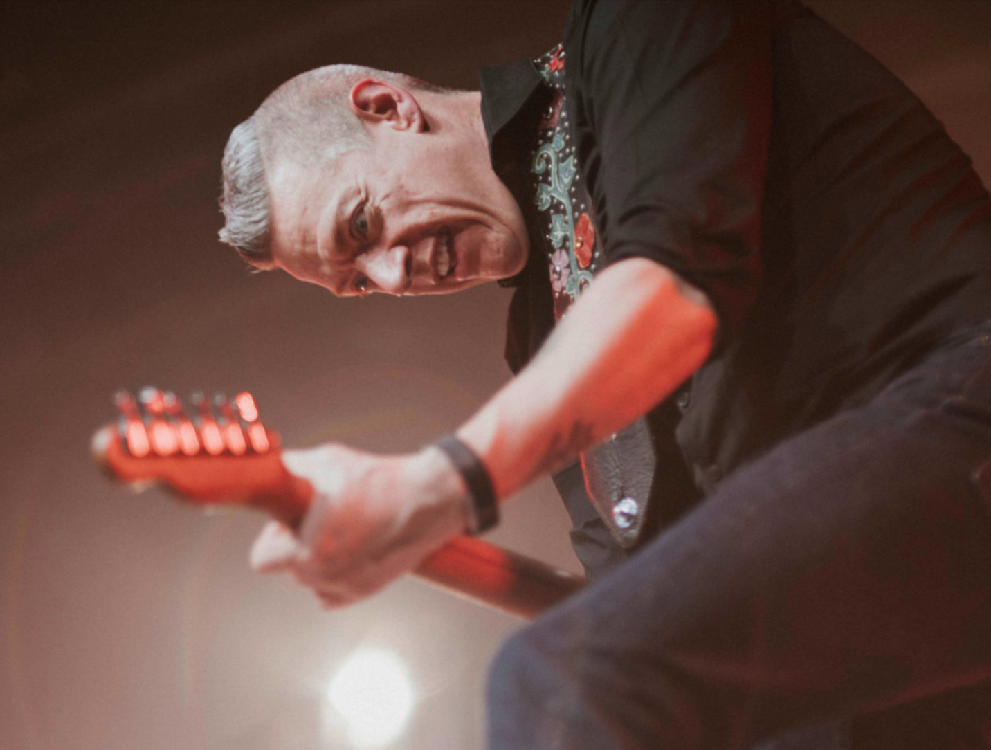 Man playing guitar, stage lit, looking down intensely; wearing black shirt, jeans.