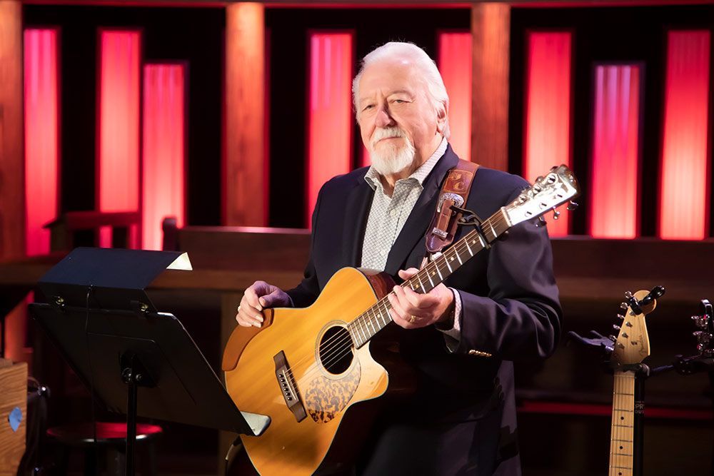 Man playing acoustic guitar in front of red-lit columns; musical score on a stand.