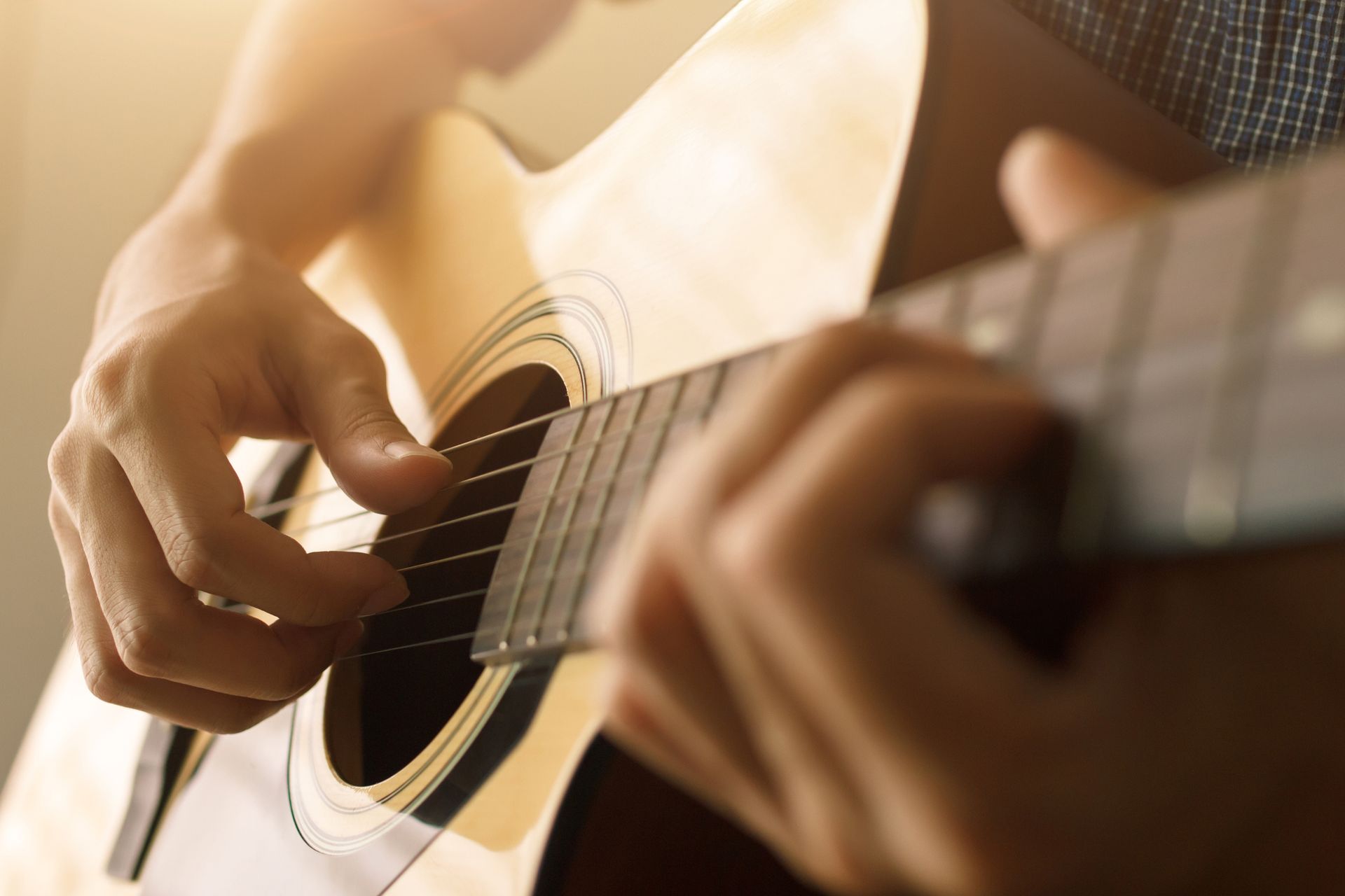 Person's hands strumming an acoustic guitar, close-up, with sunlight highlighting the wood.