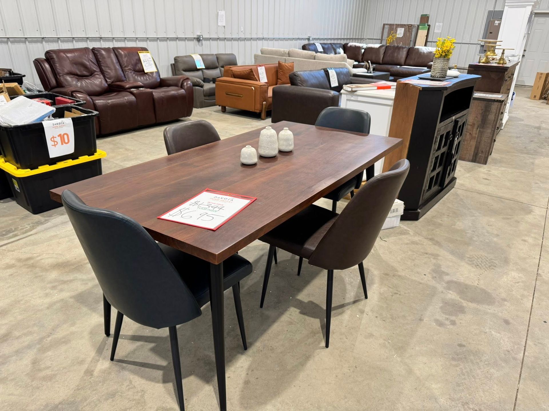 A dining table with four chairs in a warehouse. Other furniture is visible in the background.