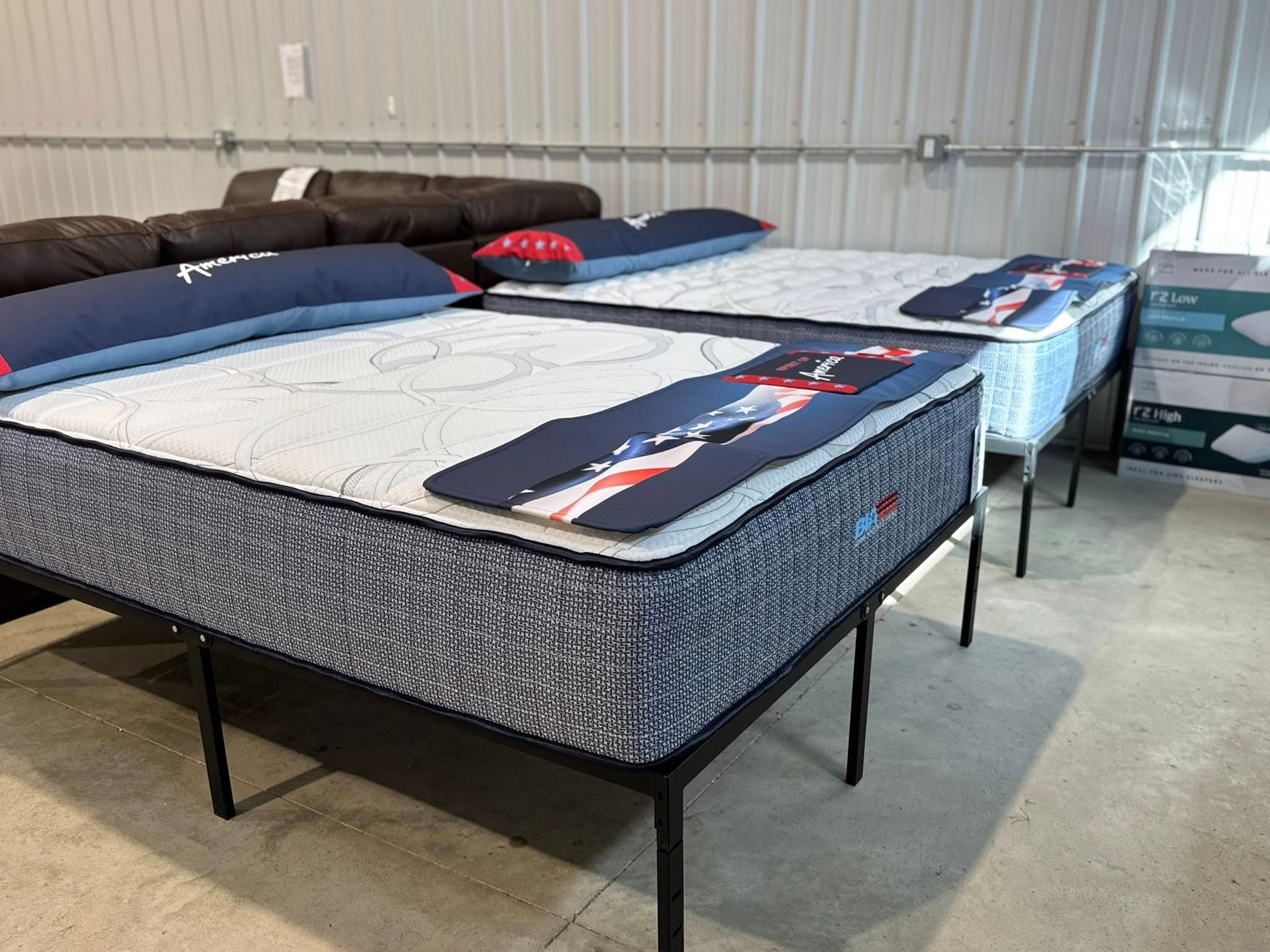 Two mattresses on black metal frames in a store. One has blue and red accents, the other has a white mattress.