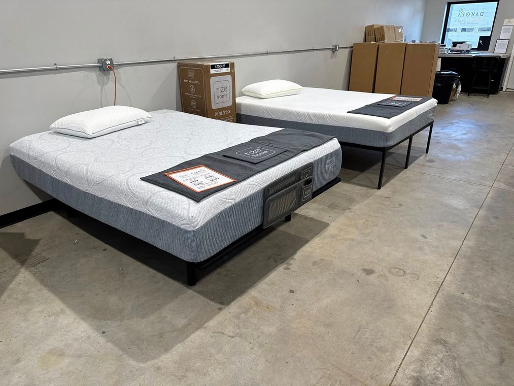 Two beds in a showroom, grey mattresses with white pillows. Boxes and other furniture are in the background.