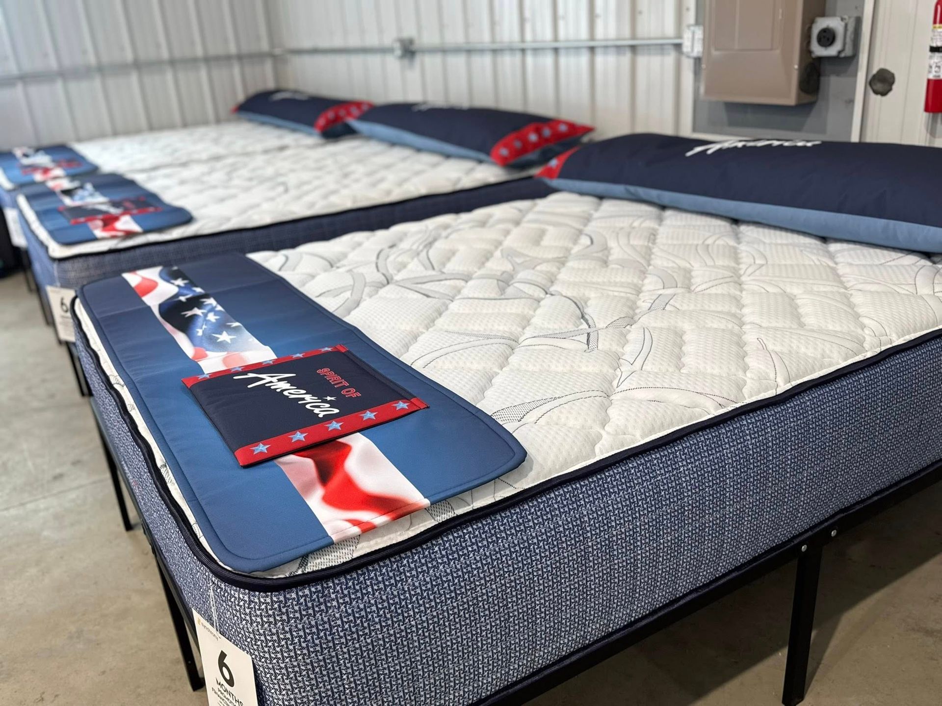 Mattresses on display in a store, featuring blue and white quilted tops, with a patriotic design on the labels.