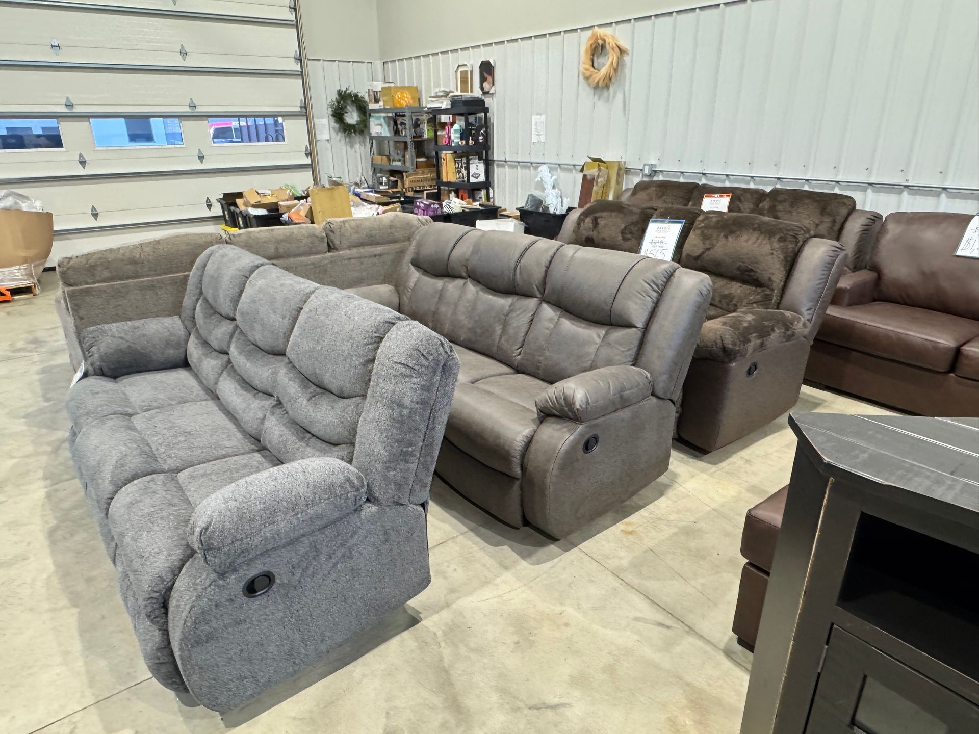 Several recliner sofas in a furniture store, various colors, some with tags, near a white wall.