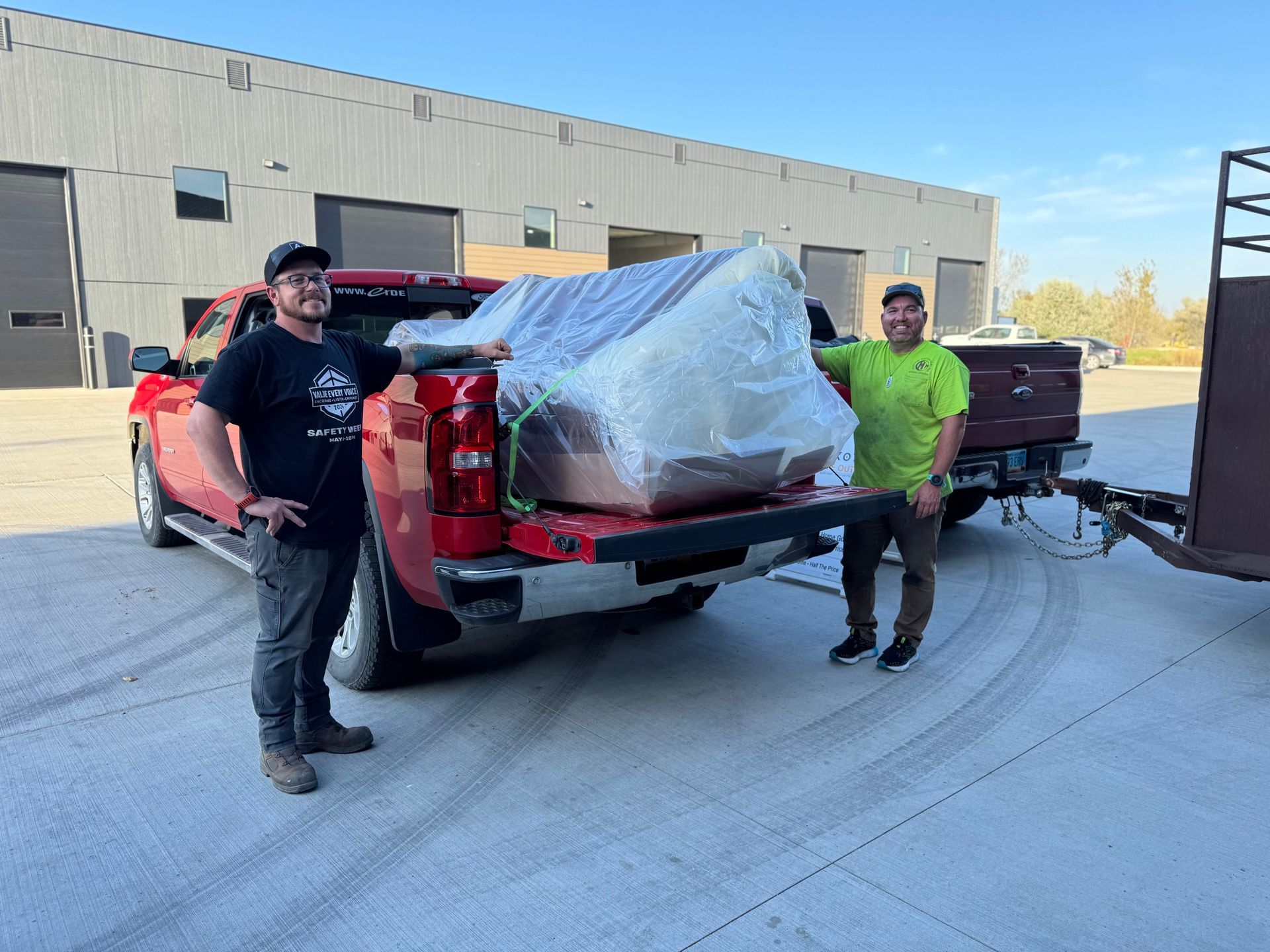 Two men stand by a red pickup truck with furniture loaded on it; outdoor setting, sunny.