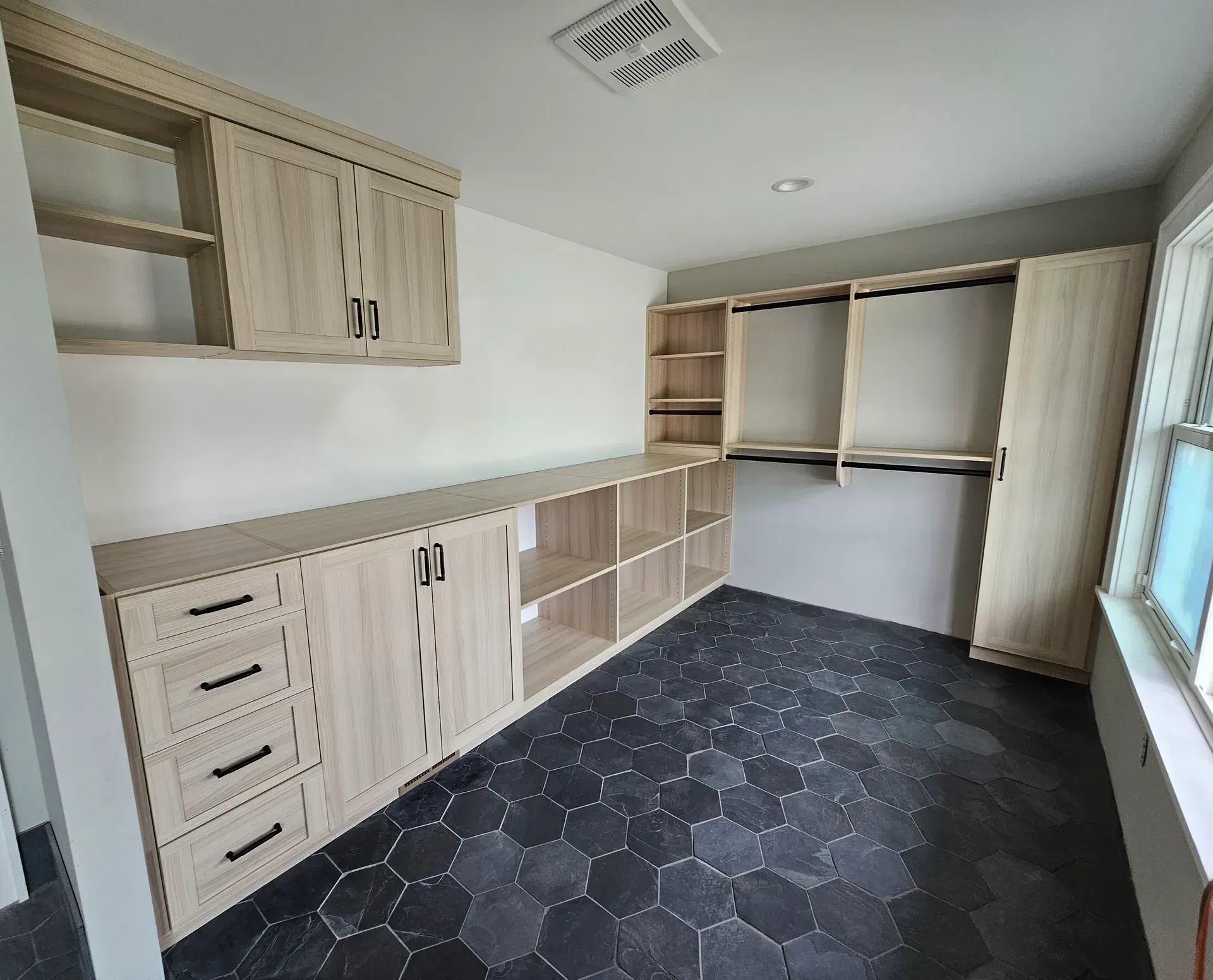 A laundry room with light wood cabinets, drawers, and black hexagonal tile flooring.
