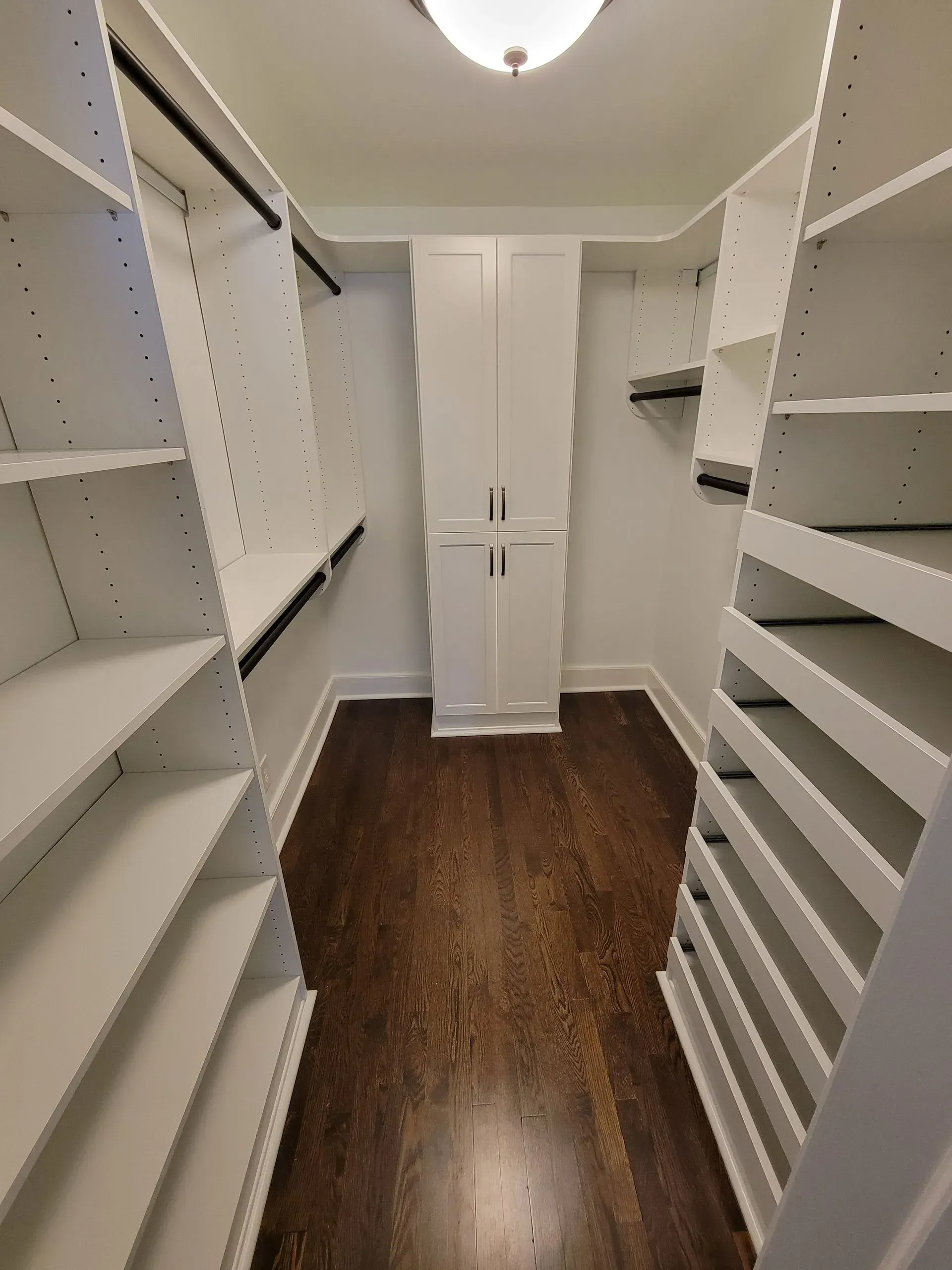 Walk-in closet with white shelves and cabinets, dark wood floor, and overhead light.