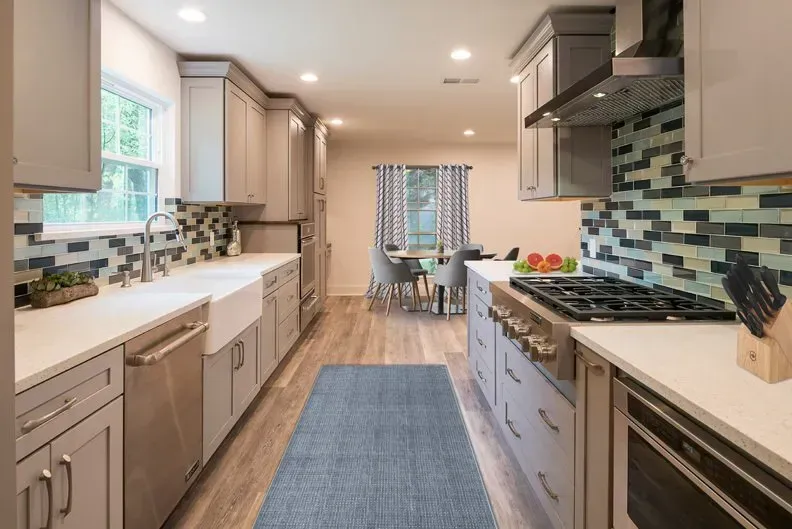 Modern kitchen with gray cabinets, white countertops, and blue backsplash. Dining area visible in the background.
